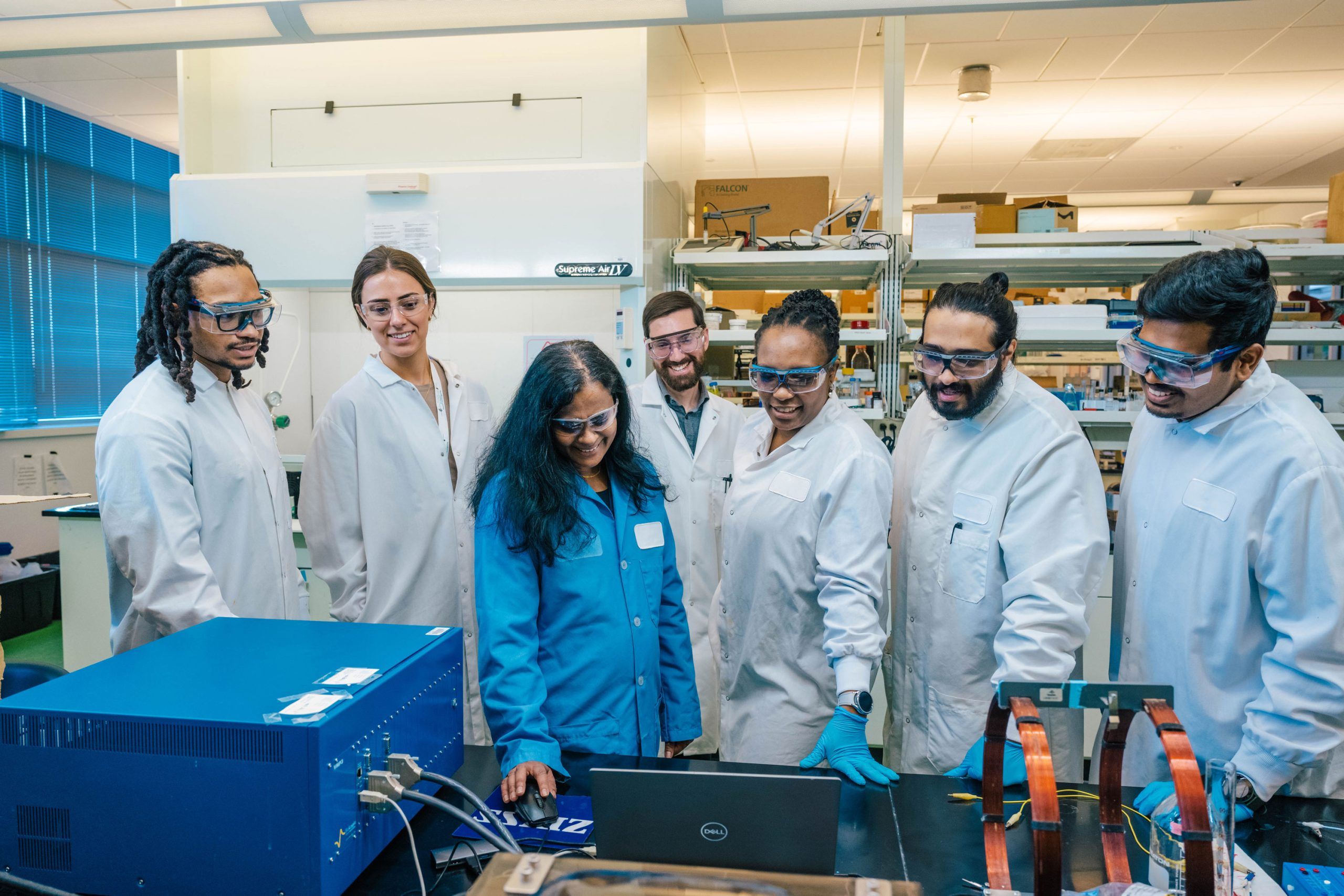 A group of seven people wearing white lab coats gather around a laboratory workstation. Dr. Rathnayake in a blue lab coat is seated in front of a laptop, while others stand closely observing. The workspace includes scientific equipment, cables, and shelves stocked with lab supplies in the background. The setting appears to be a research lab environment.