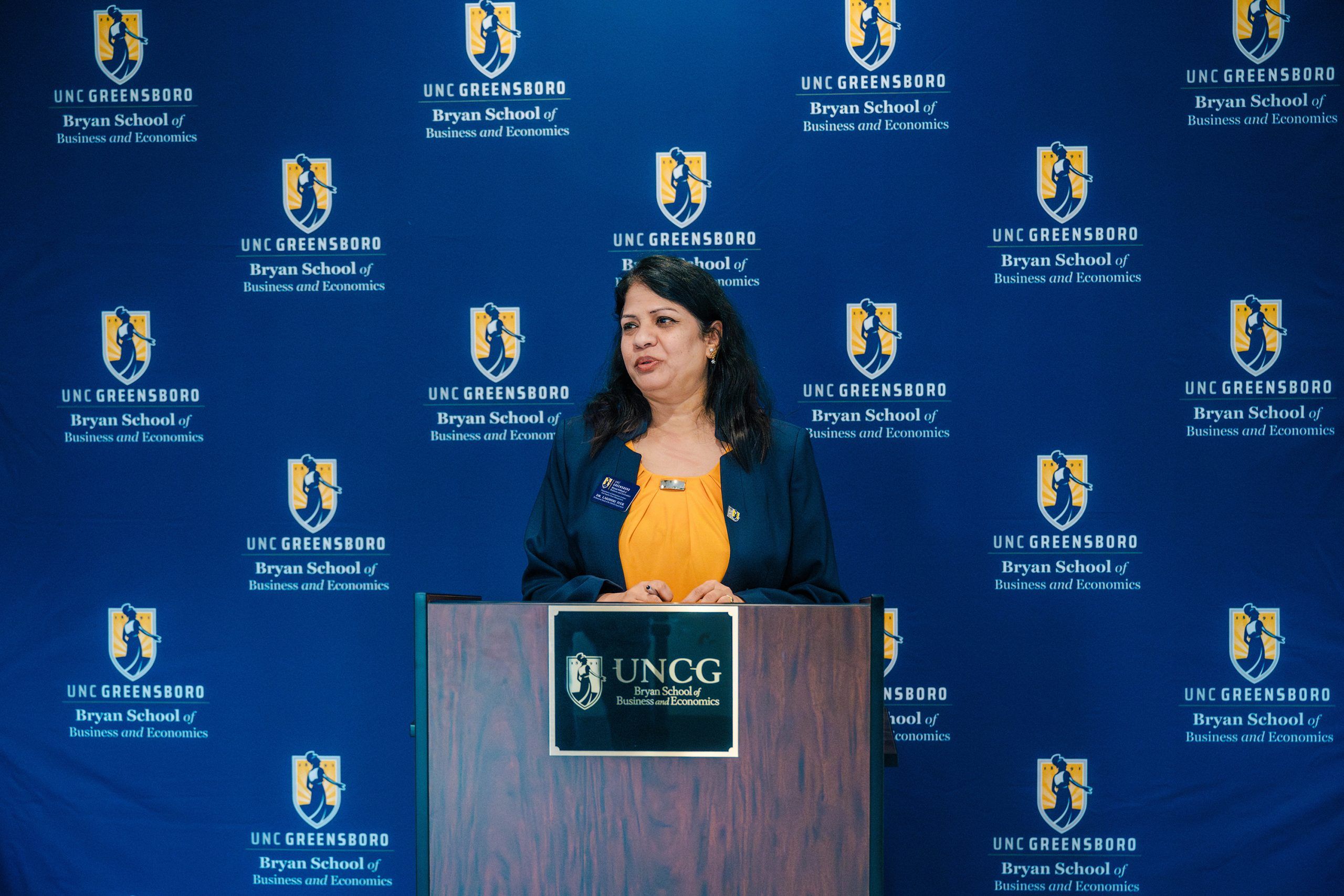 A person stands at a podium with the UNC Greensboro logo and text for the Bryan School of Business and Economics. The backdrop is a blue banner repeatedly displaying the same logo and school name.