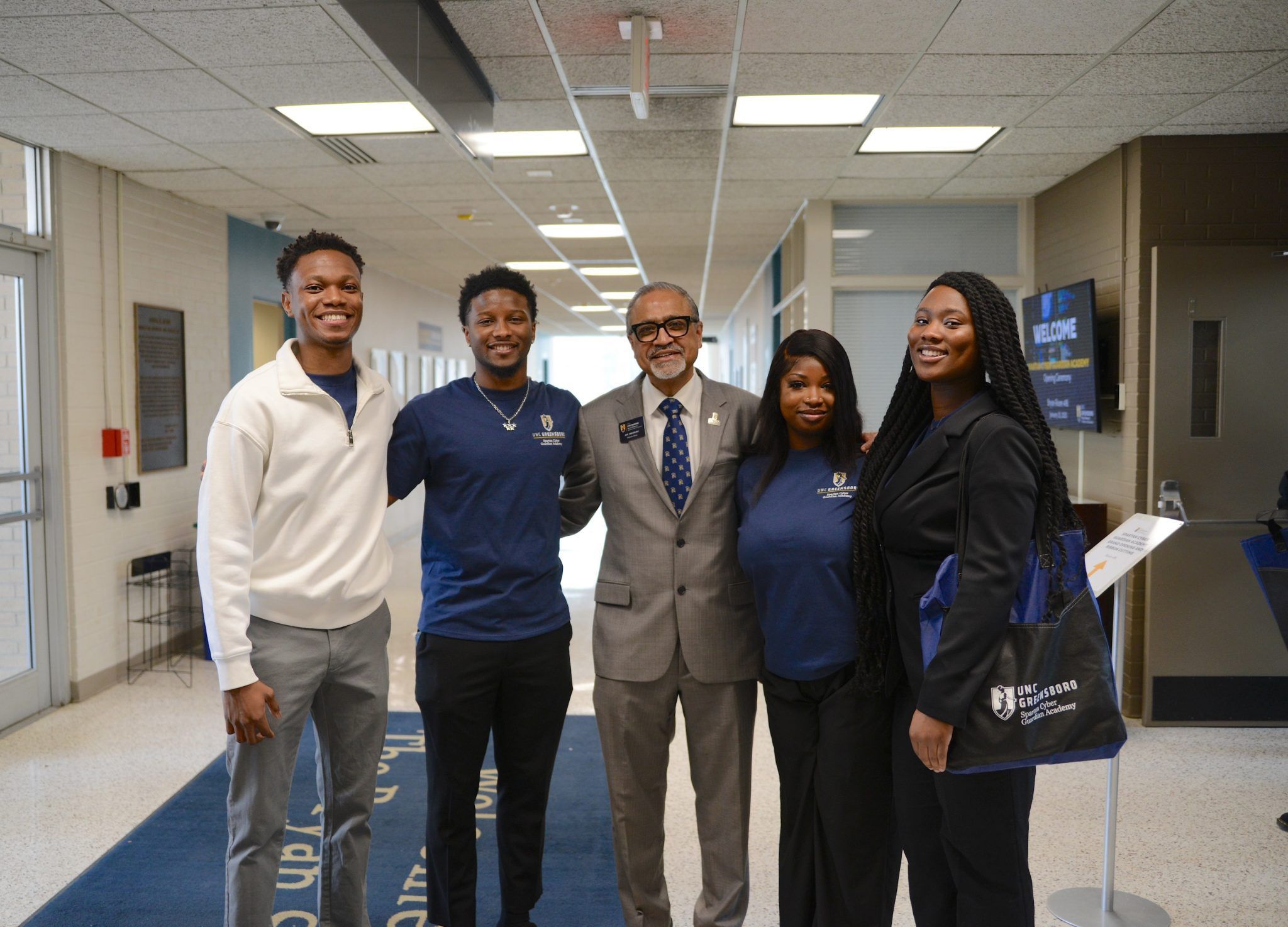 Five people stand together in a hallway, posing for a group photo. Three individuals wear navy UNC Greensboro shirts, one person is dressed in a gray suit with a name badge, and another is in a black outfit holding a UNC Greensboro tote bag. A welcome sign is visible on a monitor in the background, and the setting appears to be an event or campus building.