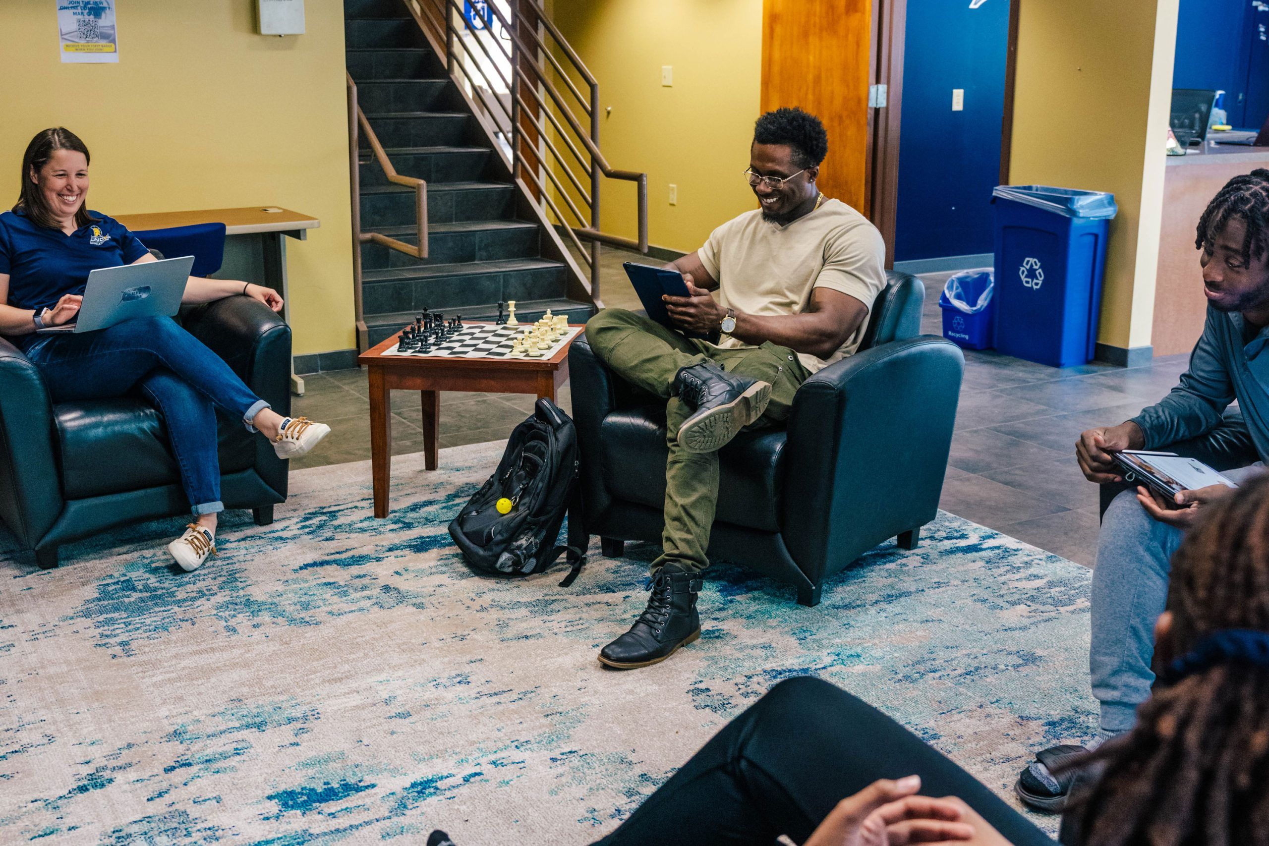 A casual meeting space with several people seated in armchairs around a small wooden table that has a chessboard on it. One person is using a laptop, another is holding a tablet, and others appear to be engaged in conversation. The setting includes a staircase in the background, a blue recycling bin, and a patterned rug on the floor.