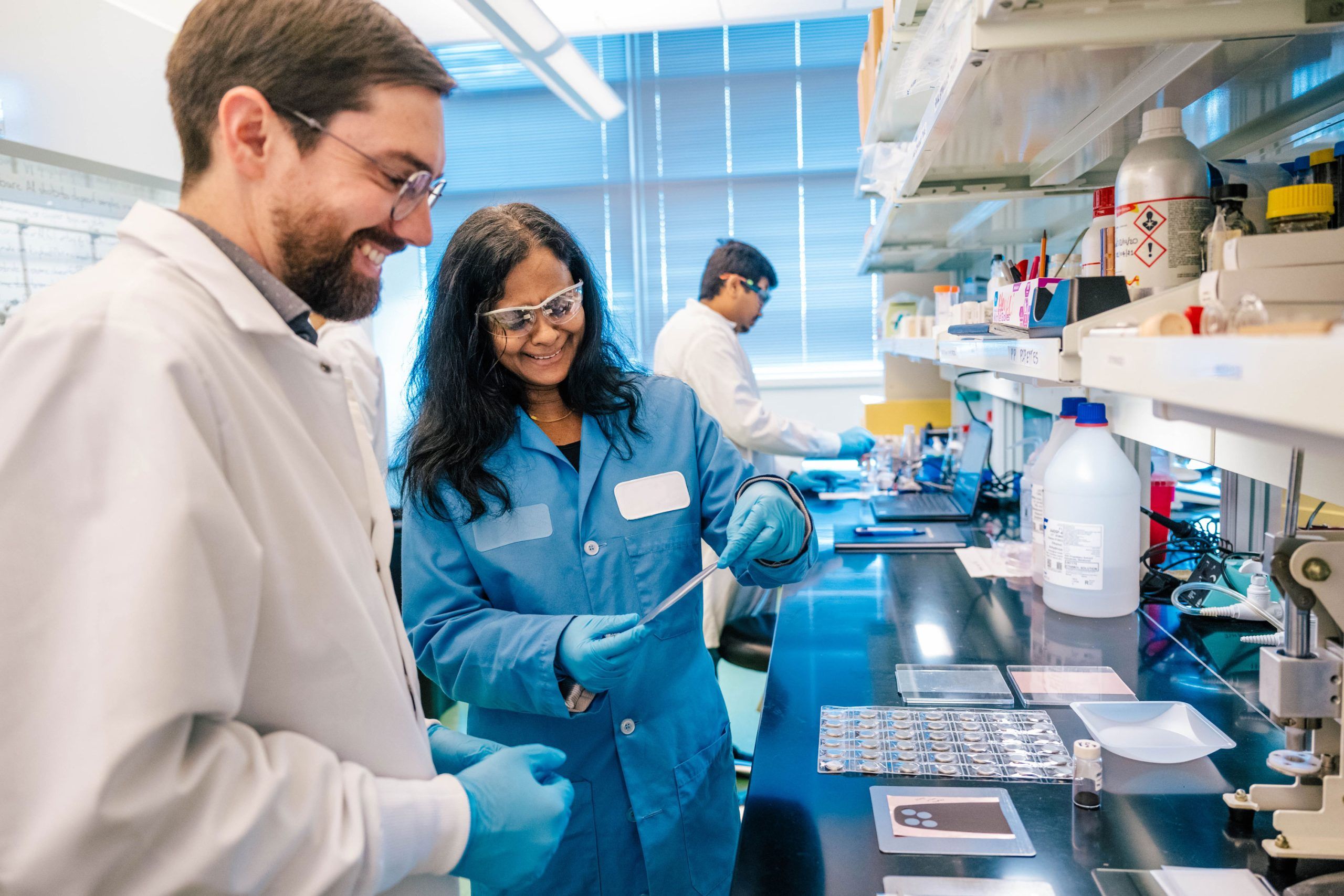 Dr. Sultana and a student wearing lab coats and blue gloves work at a laboratory bench covered with scientific equipment and materials. One person is holding a pipette and appears to be transferring liquid, while the other observes. The bench has trays with samples, a large bottle, and various lab tools. Shelves above contain containers, bottles, and supplies, and large windows in the background let in natural light.