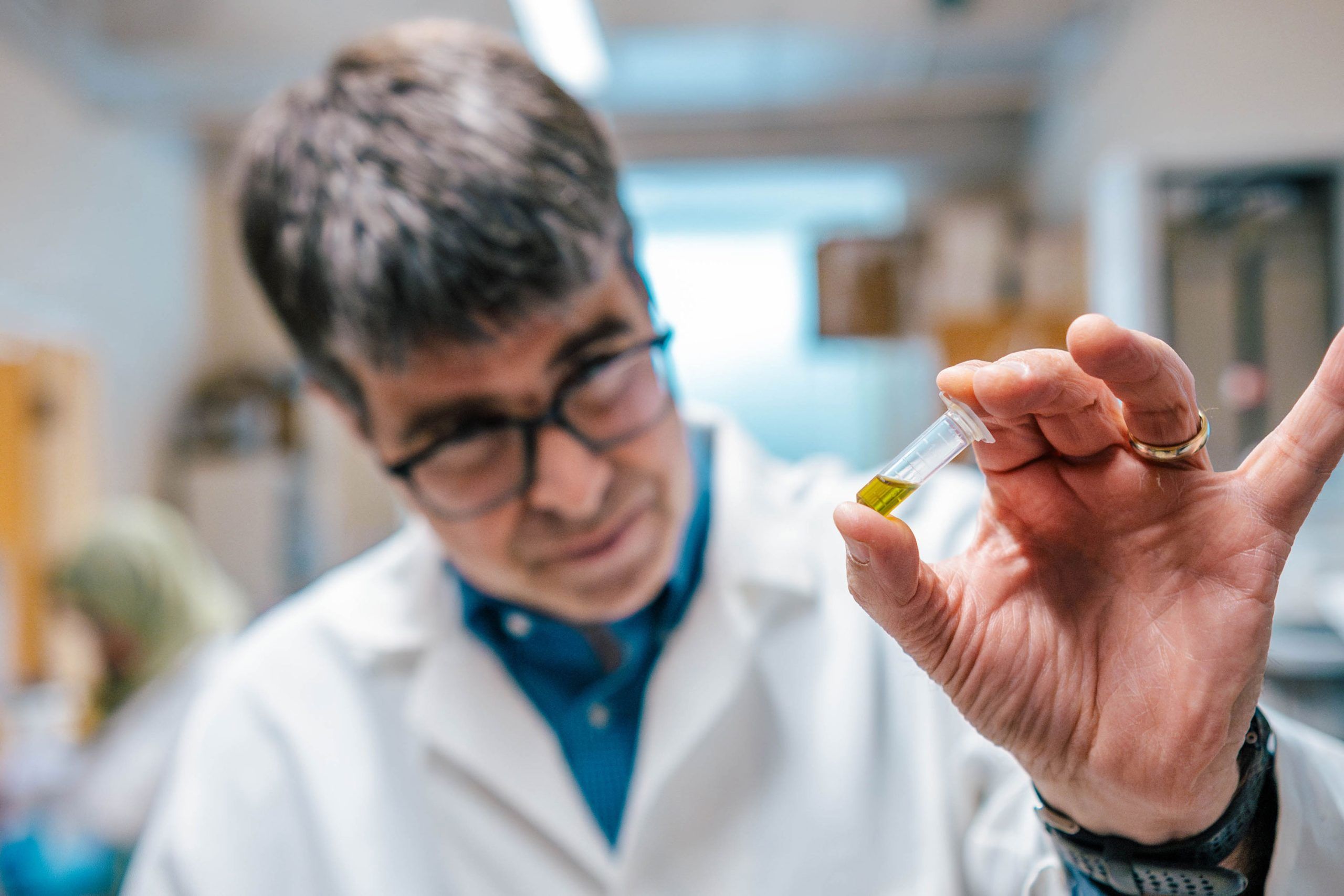 Close-up of a hand holding a small clear vial containing yellow liquid in a laboratory setting. Background shows shelves and equipment slightly out of focus.