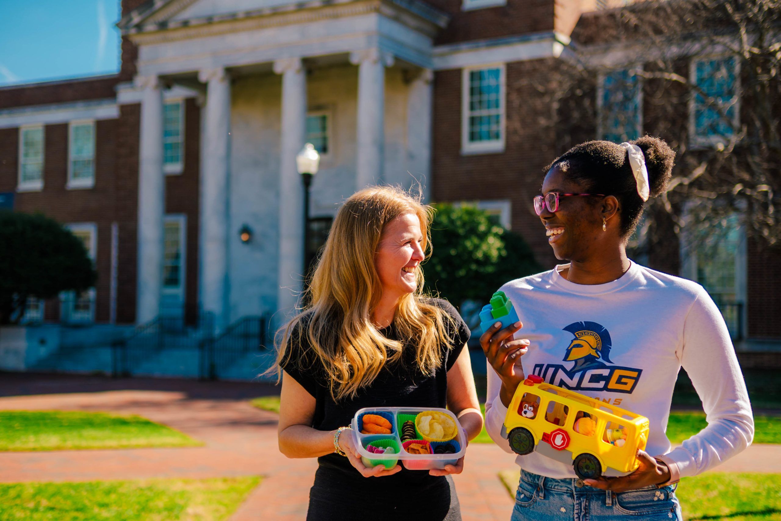 UNCG research Dr. Lenka Shriver and undergrad Jahleen Gourdine standing outside on a grassy area in front of a brick building with white columns, holding colorful educational toys including a plastic food set and a yellow toy bus.