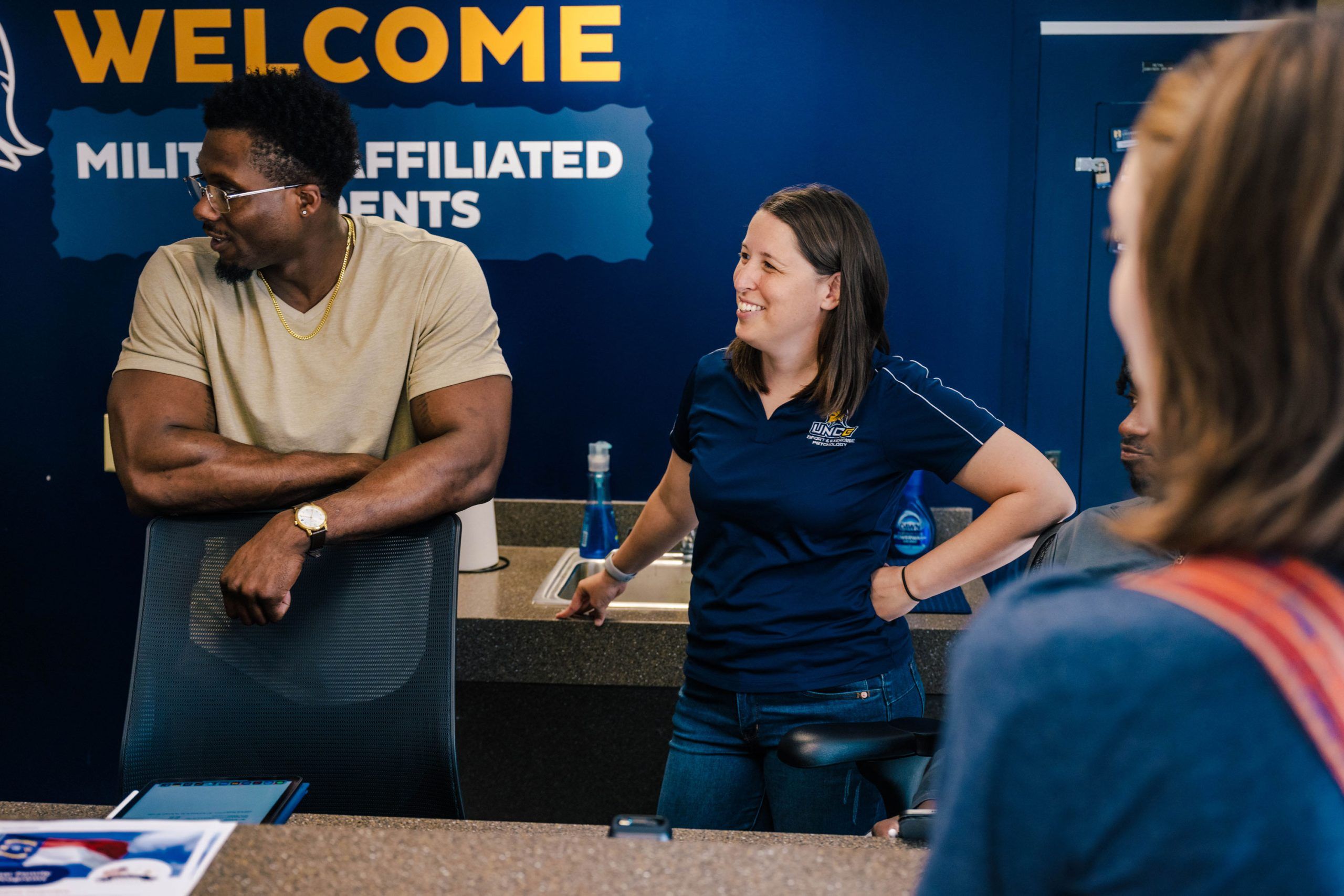 President of the UNCG chapter of the Student Veterans Association and Dr. Reifsteck stand at a counter in front of a dark blue wall with large yellow text reading “WELCOME MILITARY AFFILIATED STUDENTS.” Dr. Reifsteck is wearing a light-colored T-shirt and leaning on the counter, while the president is in a navy polo shirt with a university logo, engaged in conversation. A water bottle and informational materials are visible on the counter.