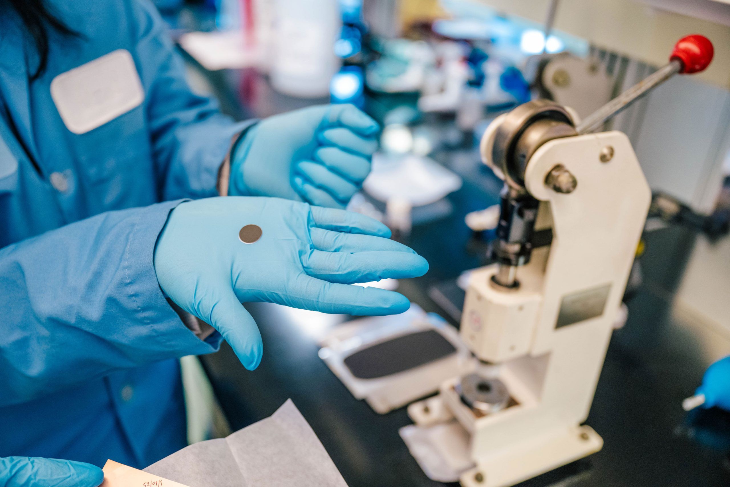 Dr. Rathnayake wearing a blue lab coat and blue gloves holds a small circular sample in one hand while standing at a laboratory bench. Nearby is a white mechanical press used for preparing materials, along with other lab equipment and tools on the work surface.