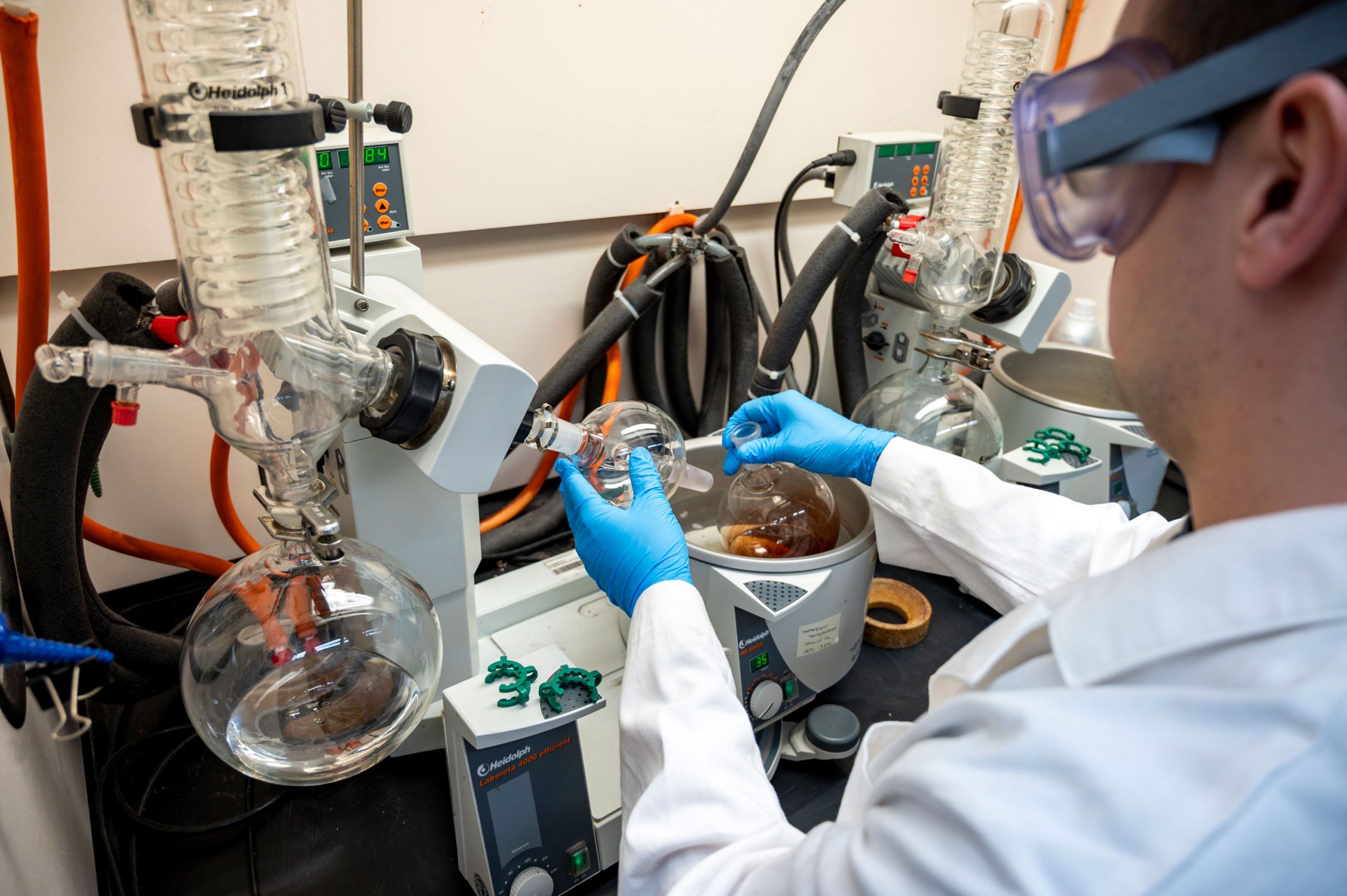 A person wearing a white lab coat and blue gloves operates a rotary evaporator in a laboratory. The setup includes large round-bottom glass flasks connected to a condenser and tubing, with one flask partially filled with liquid. The equipment is mounted on a benchtop alongside other lab instruments and control panels, and the background shows additional hoses and safety features typical of a chemistry lab.