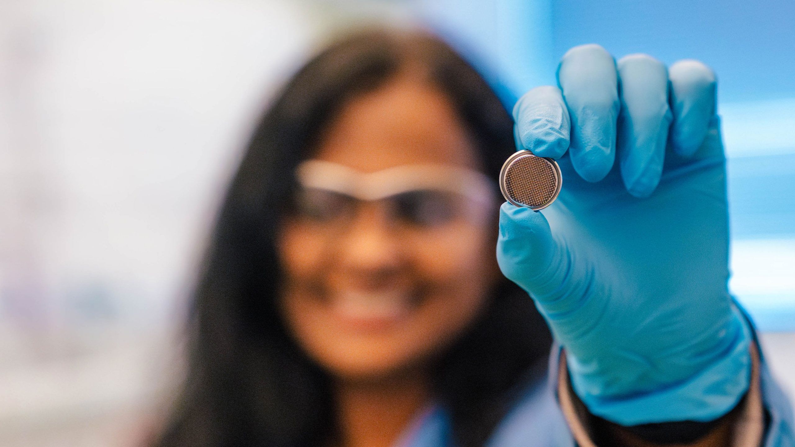 A gloved hand holds a small, round metallic object resembling a coin cell battery or sensor component. The object has a textured or mesh-like surface on one side. The background is softly blurred, showing a laboratory setting with cool blue tones.