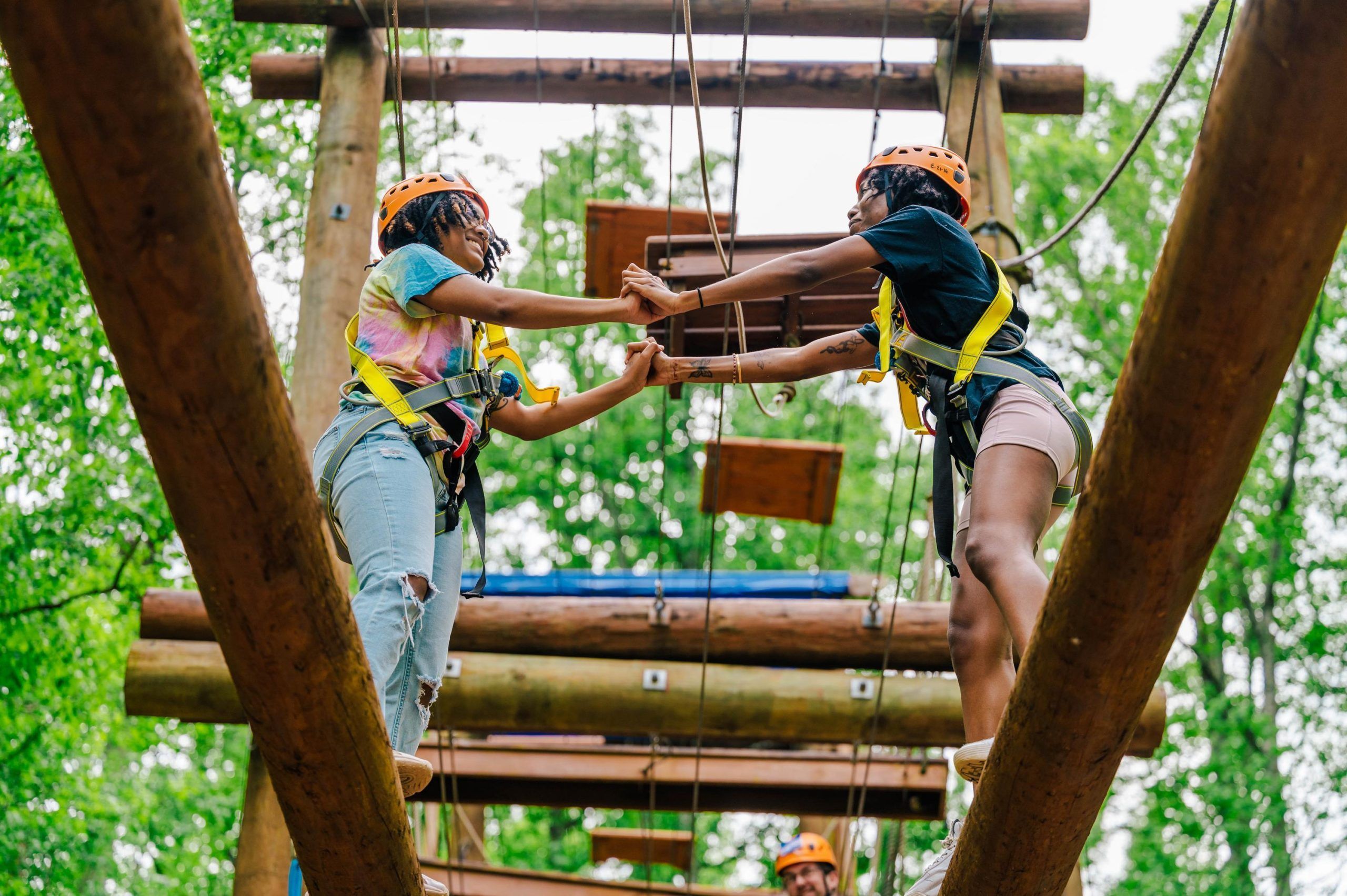 Two people wearing orange helmets and safety harnesses balance on wooden beams during a ropes course activity. They are holding hands for support while navigating the elevated structure, which includes ropes and wooden platforms. Green trees fill the background, indicating an outdoor adventure setting.