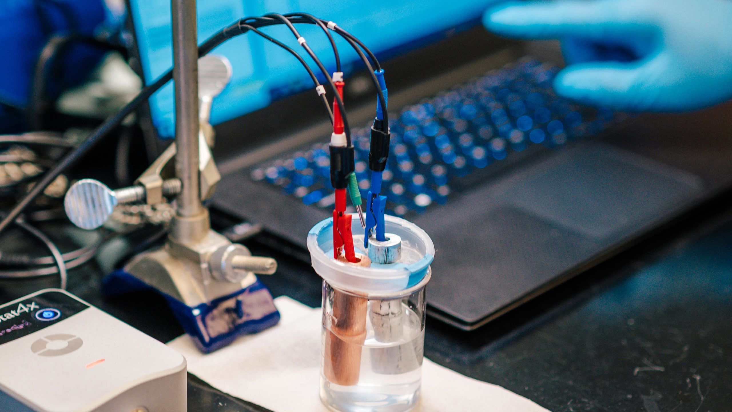 A close-up view of a laboratory setup showing an electrochemical experiment. A small container filled with clear liquid holds two metal electrodes connected to red, blue, and black wires, which are attached to a stand. The wires lead to a nearby device and a laptop in the background. A gloved hand is visible near the laptop, suggesting data monitoring or adjustment during the experiment.