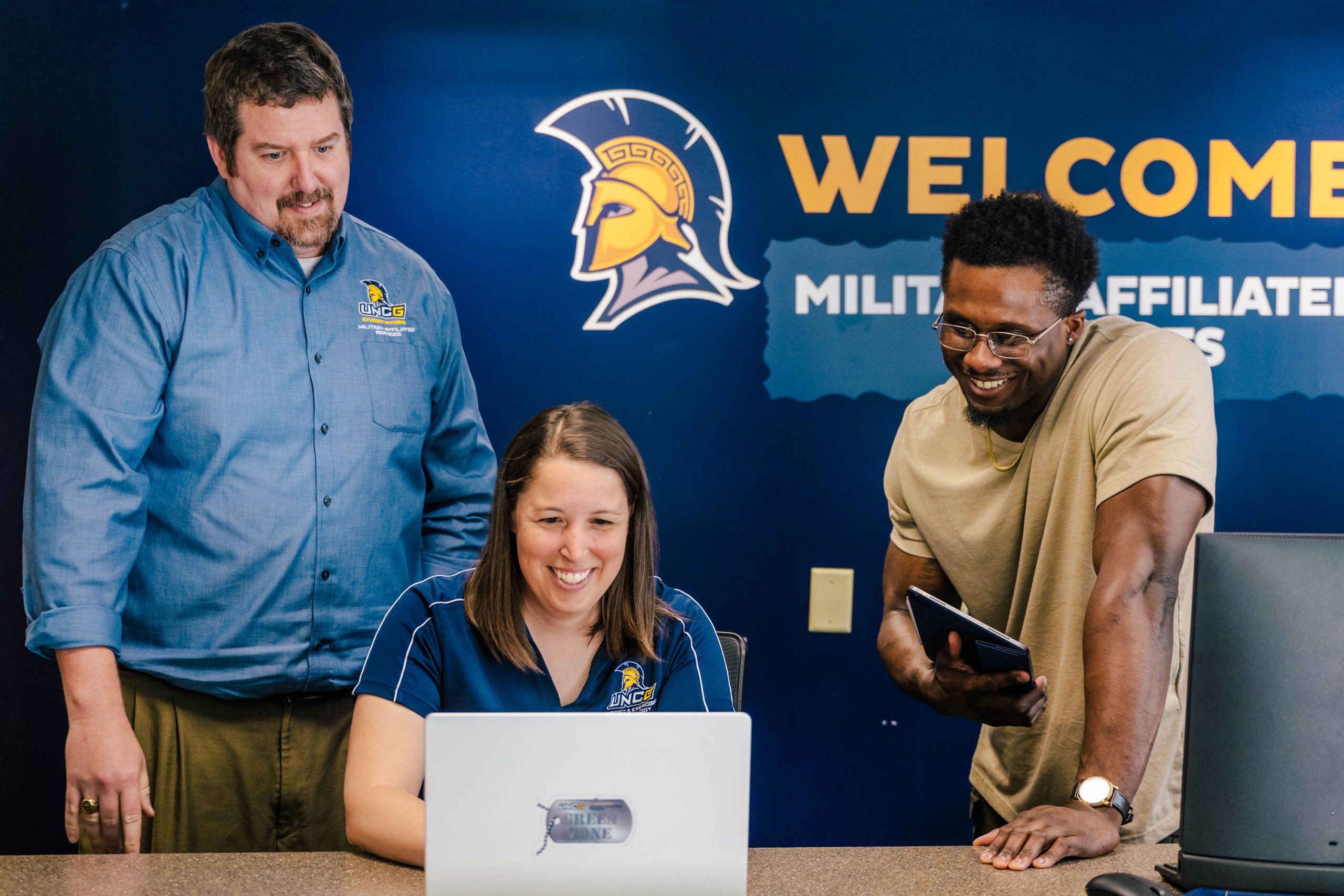 Gregory, Reifsteck, and the president of the UNCG chapter of the Student Veterans Association are gathered at a desk in front of a dark blue wall with the UNC Greensboro Spartan logo and the words “WELCOME MILITARY AFFILIATED STUDENTS.” One person is seated working on a laptop, while another stands beside holding a tablet, and the third stands on the opposite side wearing a UNC Greensboro shirt. The setting appears to be an office or resource center.