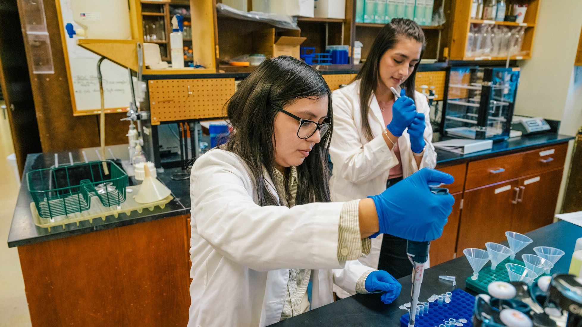 Selena Villa and an undergraduate nutrition student wearing lab coats and blue gloves working at a laboratory bench; the undergrad student is using a pipette to transfer liquid into small tubes, with racks, funnels, and other lab equipment visible on the counter.