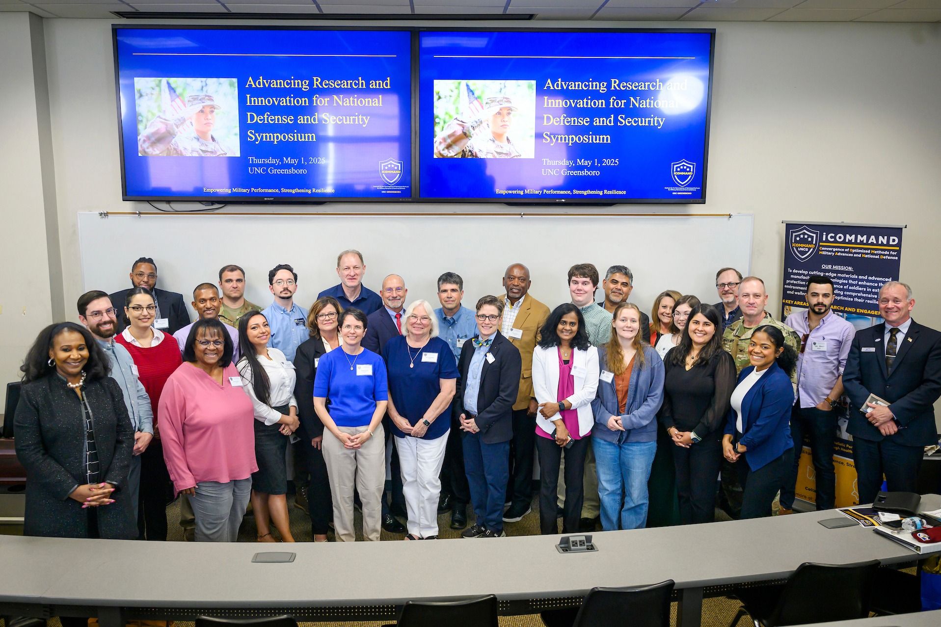 A large group of people pose for a photo in a lecture hall in front of two screens displaying the text “Advancing Research and Innovation for National Defense and Security Symposium” along with the UNC Greensboro logo. The group stands in front of a whiteboard, and the room has tiered seating with desks and microphones visible in the foreground.