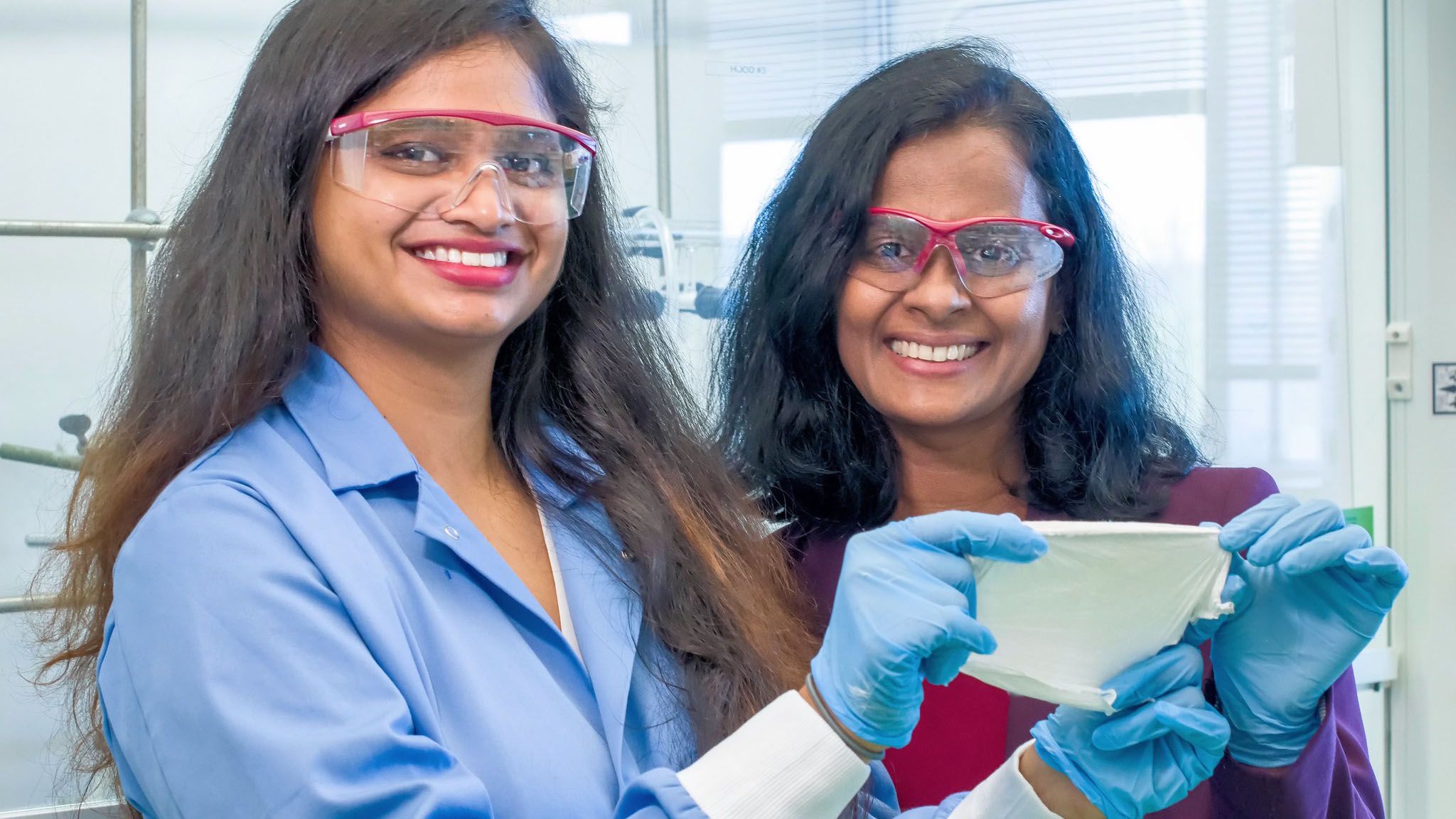 Dr. Rathnayake and her student stand in a laboratory wearing gloves and safety glasses, examining a white filter or membrane