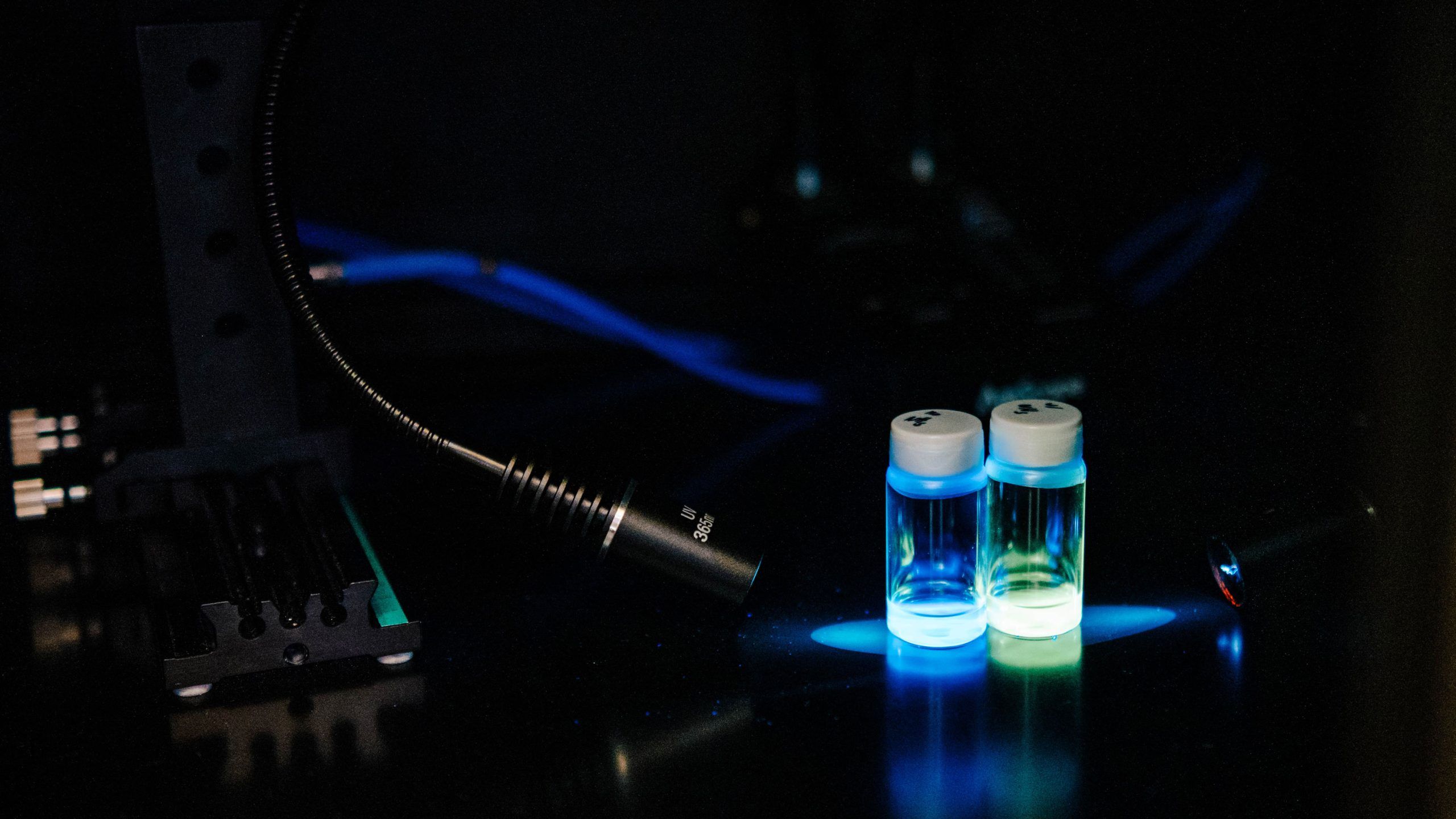Two small glass vials with white caps sit on a dark surface, illuminated by a bright blue and green glow from underneath. The vials contain clear liquid that reflects the colored light. Nearby, a flexible black lamp and blue cables are visible, along with parts of scientific equipment, suggesting a laboratory setting focused on optical or fluorescence experiments.