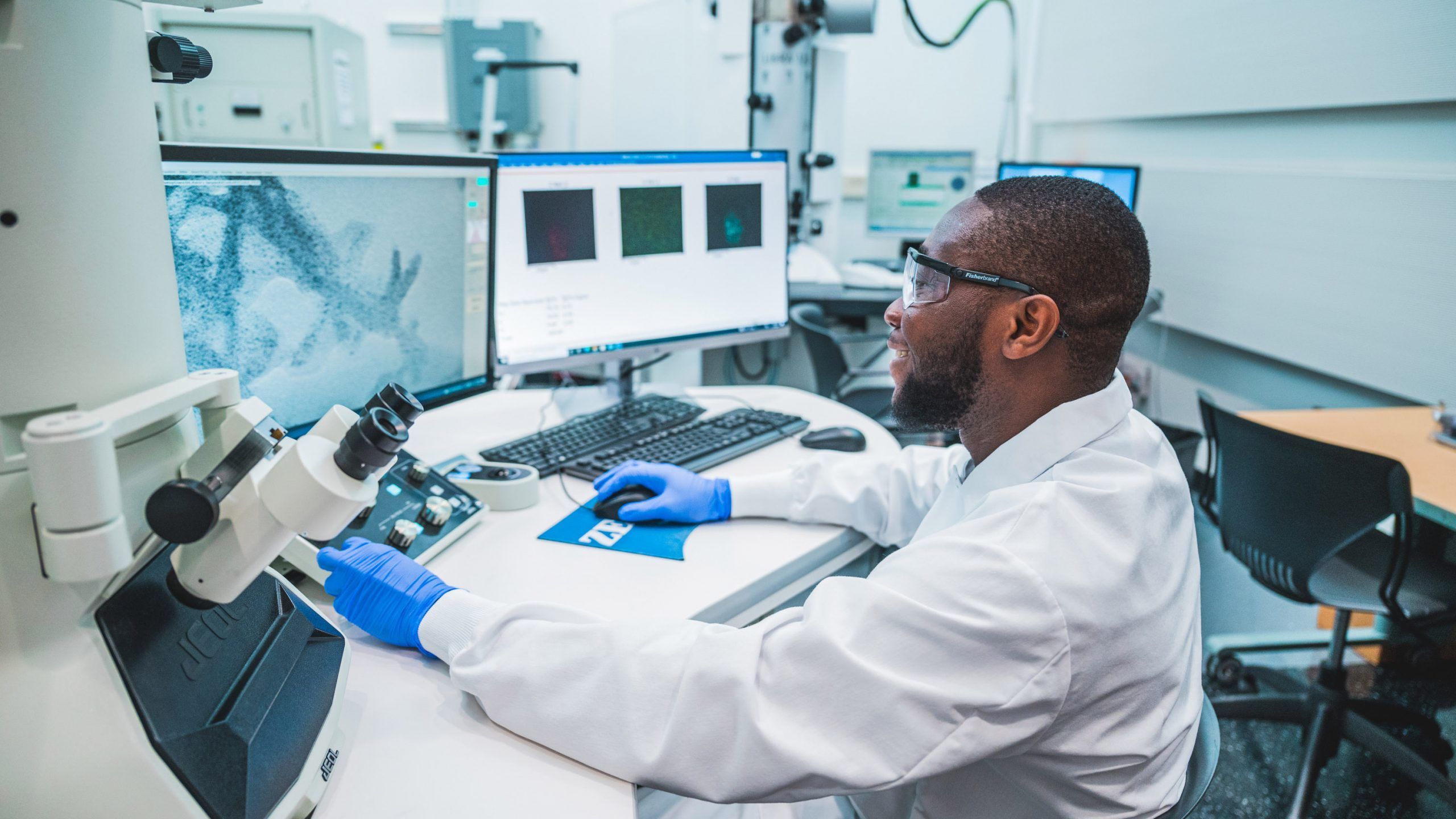 Kelvin Adrah, a PhD nanoscience student, wears a lab coat while working on a computer in a lab. 