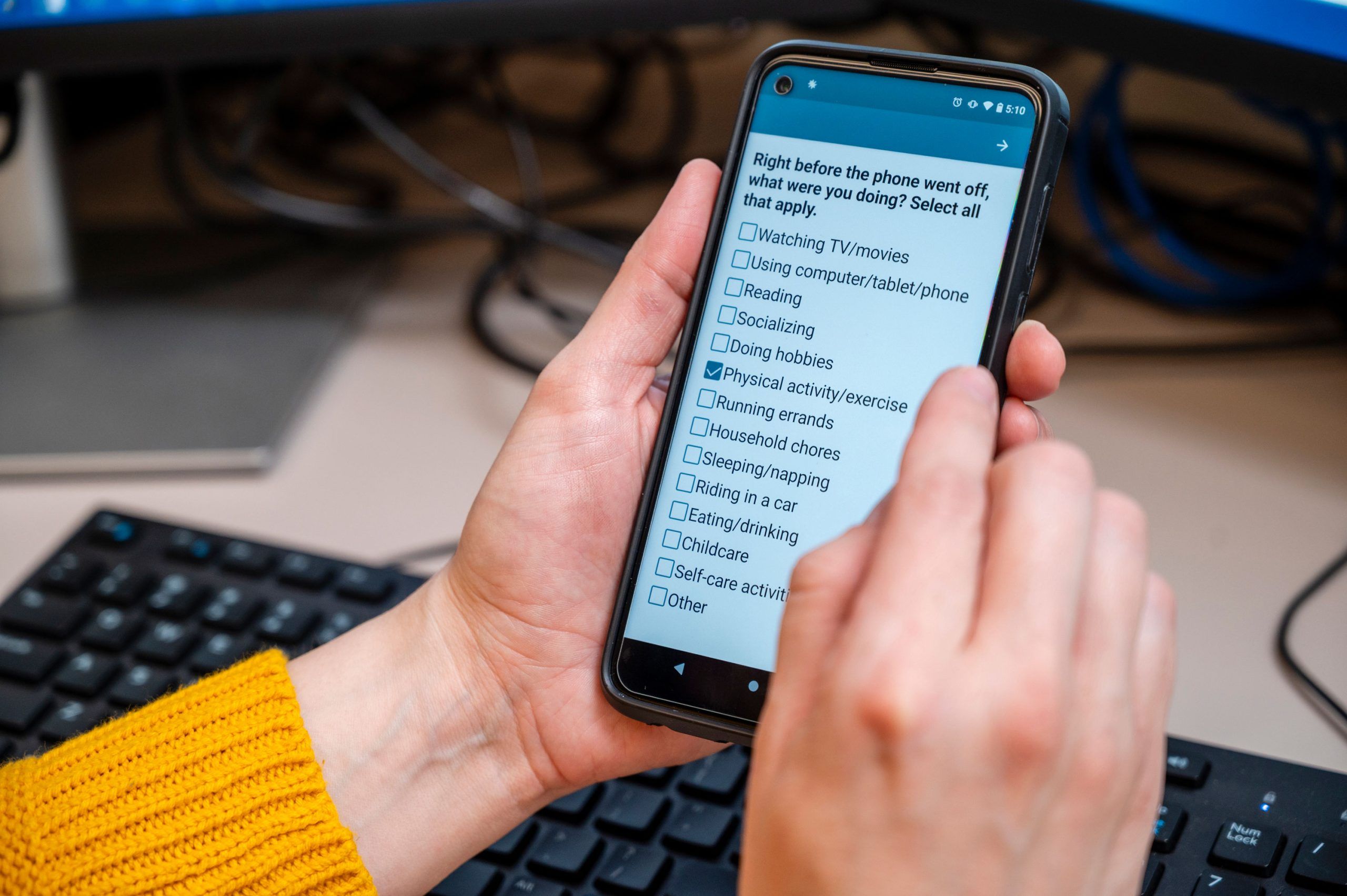 Dr. Maher holds a smartphone displaying a survey question that reads, “Right before the phone went off, what were you doing? Select all that apply.” The screen shows a checklist with options such as watching TV/movies, reading on a computer/tablet/phone, socializing, doing hobbies, physical activity/exercise, and others. A keyboard and part of a desk are visible in the background.