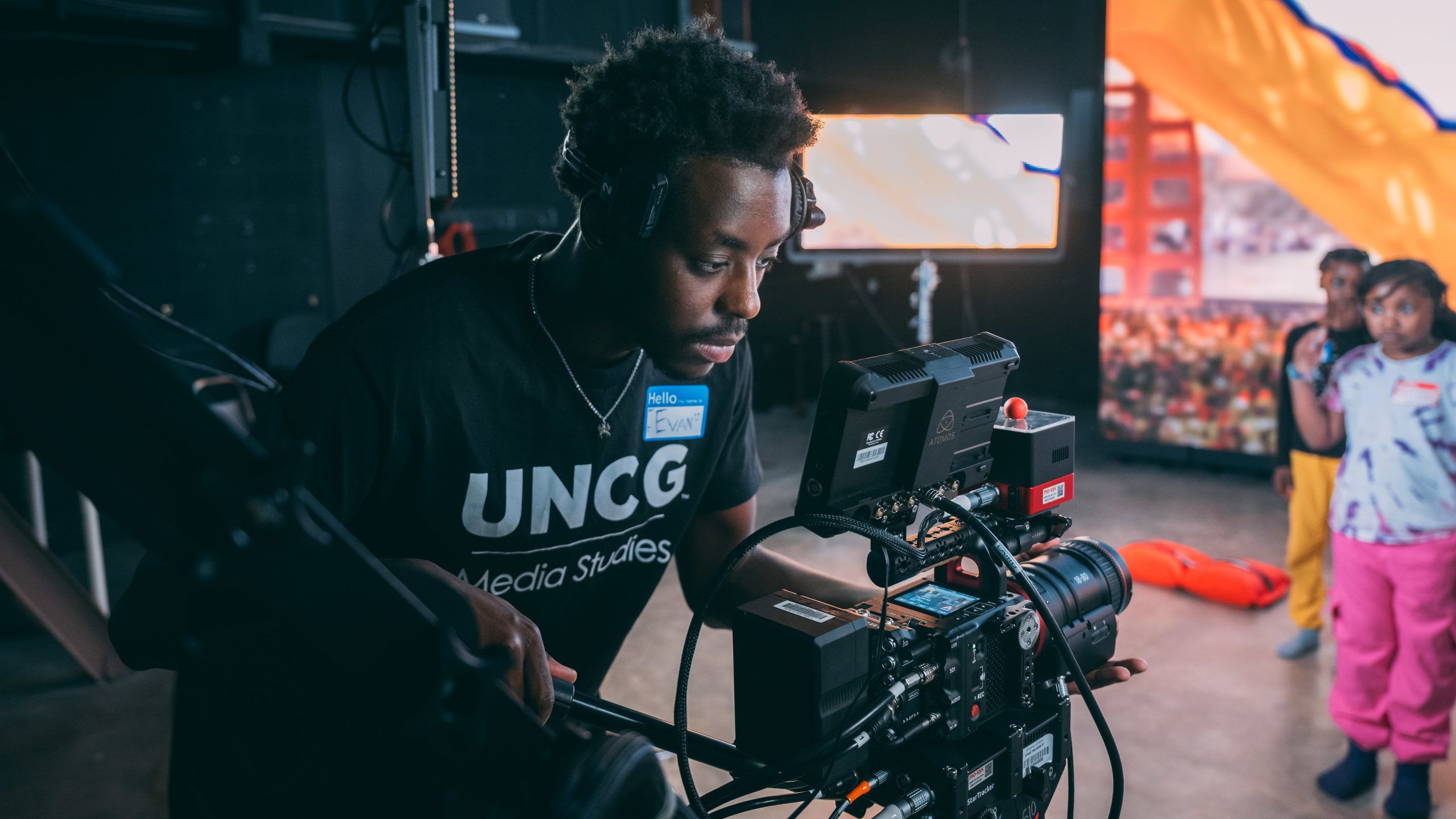 A person wearing a black T-shirt with “UNCG Media Studies” printed on it operates a professional video camera mounted on a rig. The camera has multiple attachments, including a monitor and cables. The setting appears to be an indoor studio or event space with dim lighting, a large orange fabric backdrop, and a screen displaying visuals in the background. Two other individuals stand further back near the backdrop.