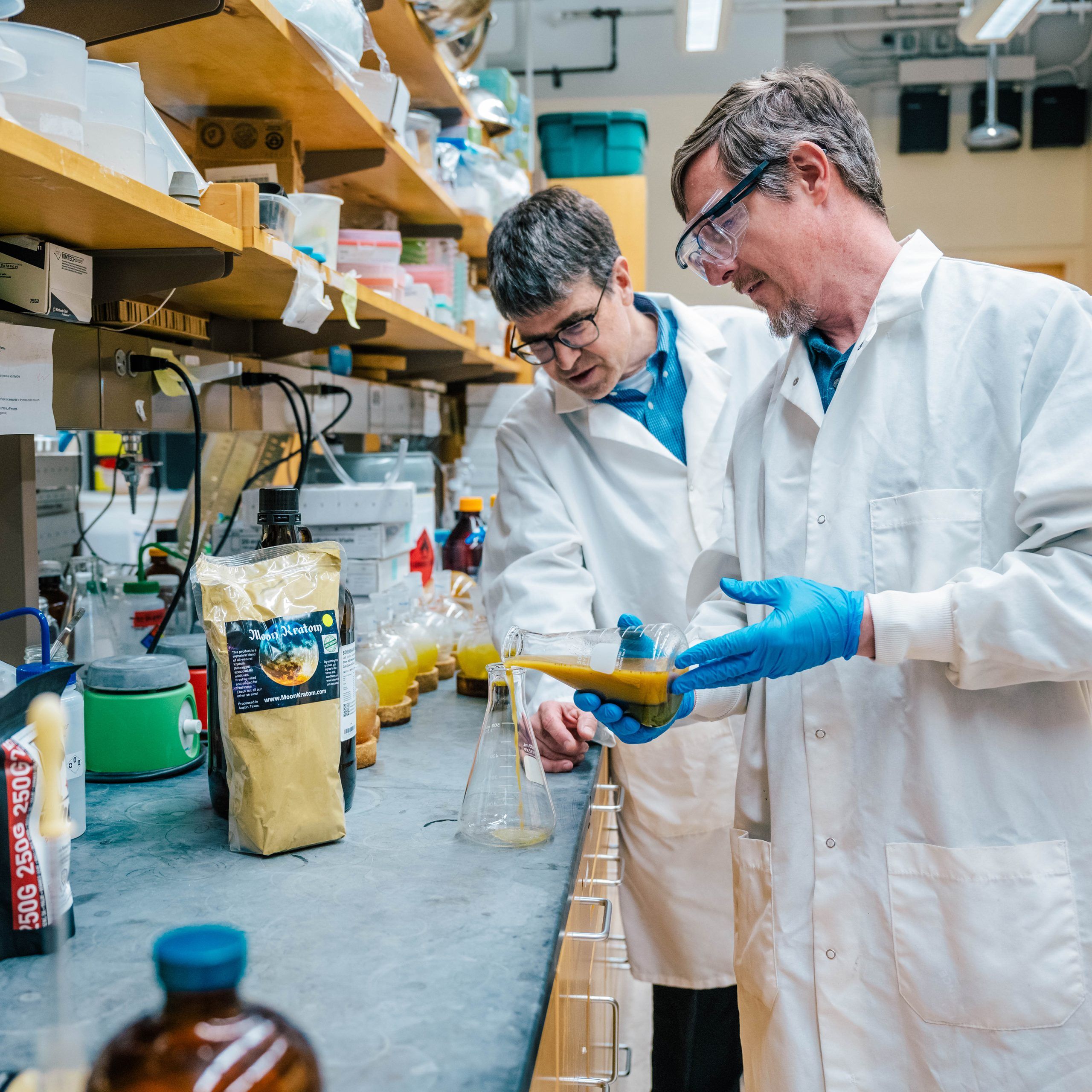 Dr. Oberlies wearing a white lab coat and blue gloves working at a laboratory bench filled with scientific equipment and containers. One person is pouring a yellow liquid from a flask into another vessel, while various bottles, bags of powdered substances, and glassware are arranged on the countertop. Shelves above hold additional supplies, and the background shows typical lab fixtures and equipment.