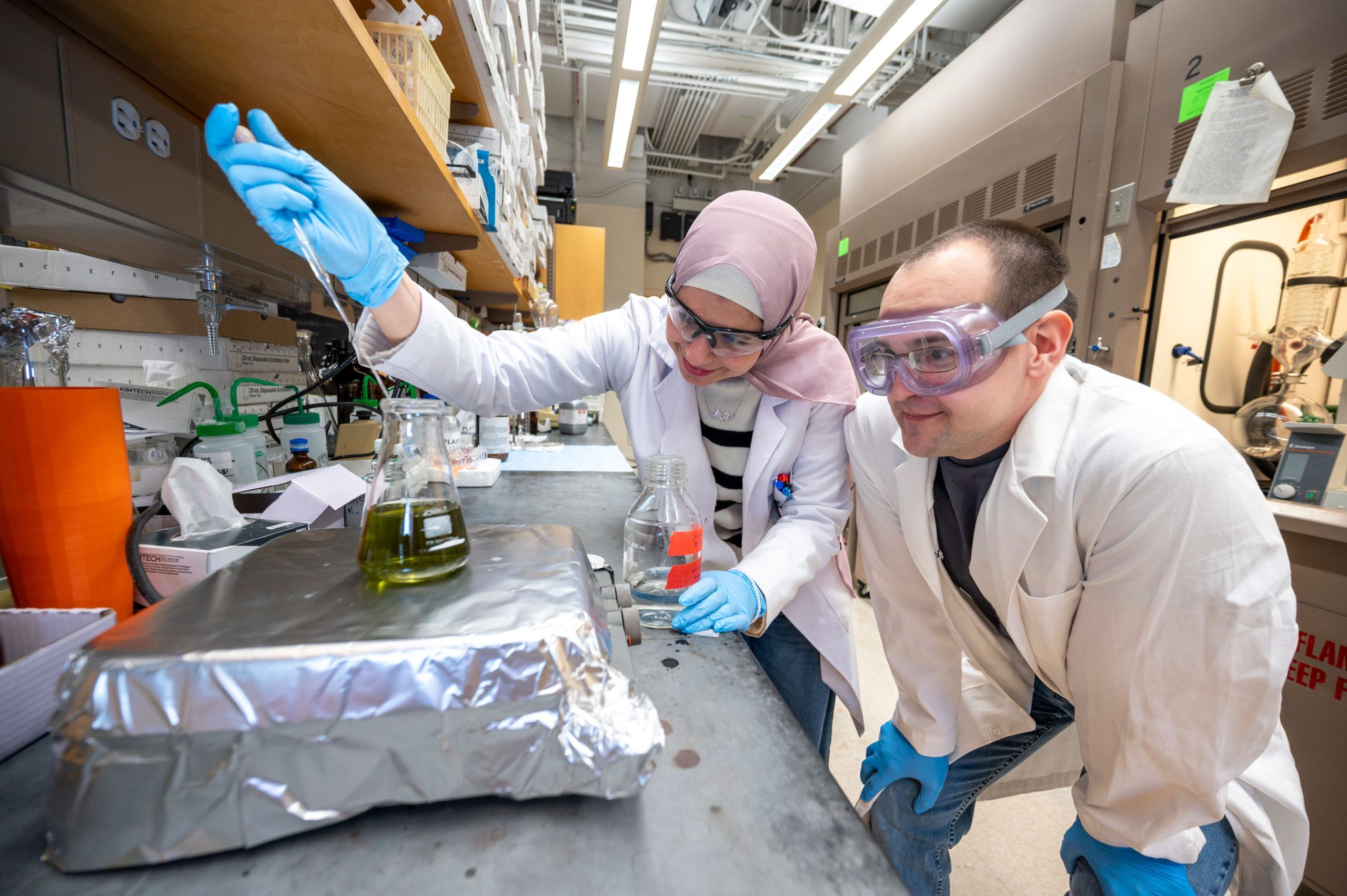 Doctoral Student Reema Al-Qiam and a student wearing white lab coats and blue gloves work together at a laboratory bench. One person is using a metal tool to manipulate or stir a substance in a glass beaker containing a green liquid, which is placed on a surface covered with foil. Another clear glass bottle is nearby, along with various lab supplies and equipment on the counter. Shelves above hold containers and materials, and the background shows a fume hood and other standard lab fixtures.
