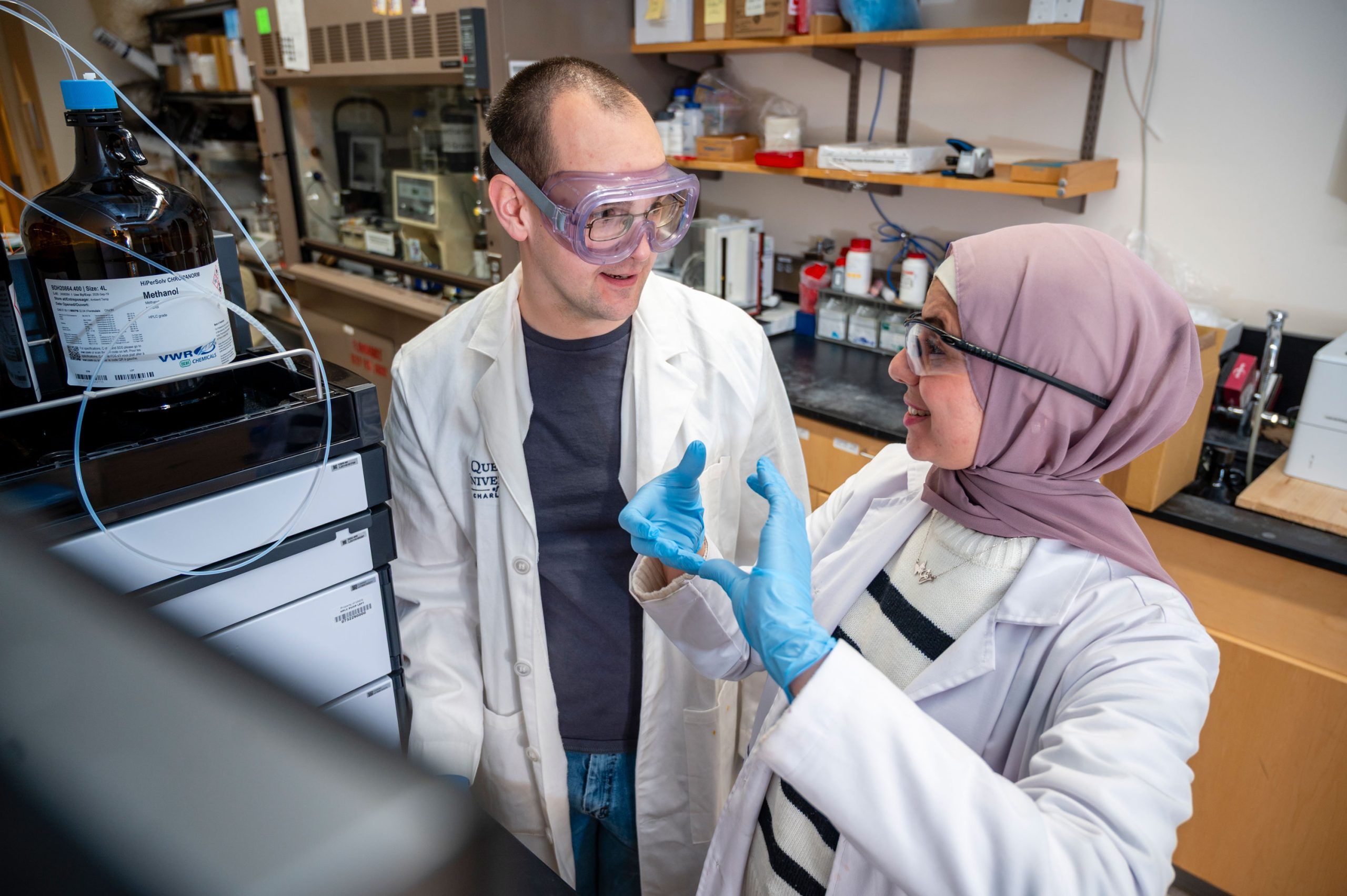 Doctoral student Al-Qiam and a student wearing white lab coats stand in a laboratory near a large analytical instrument with tubing connected to a dark glass bottle labeled “Methanol.” One person is gesturing with gloved hands as if explaining a process, while the other observes. The background includes shelves with various containers, lab equipment, and a countertop with instruments, creating a typical research lab environment.