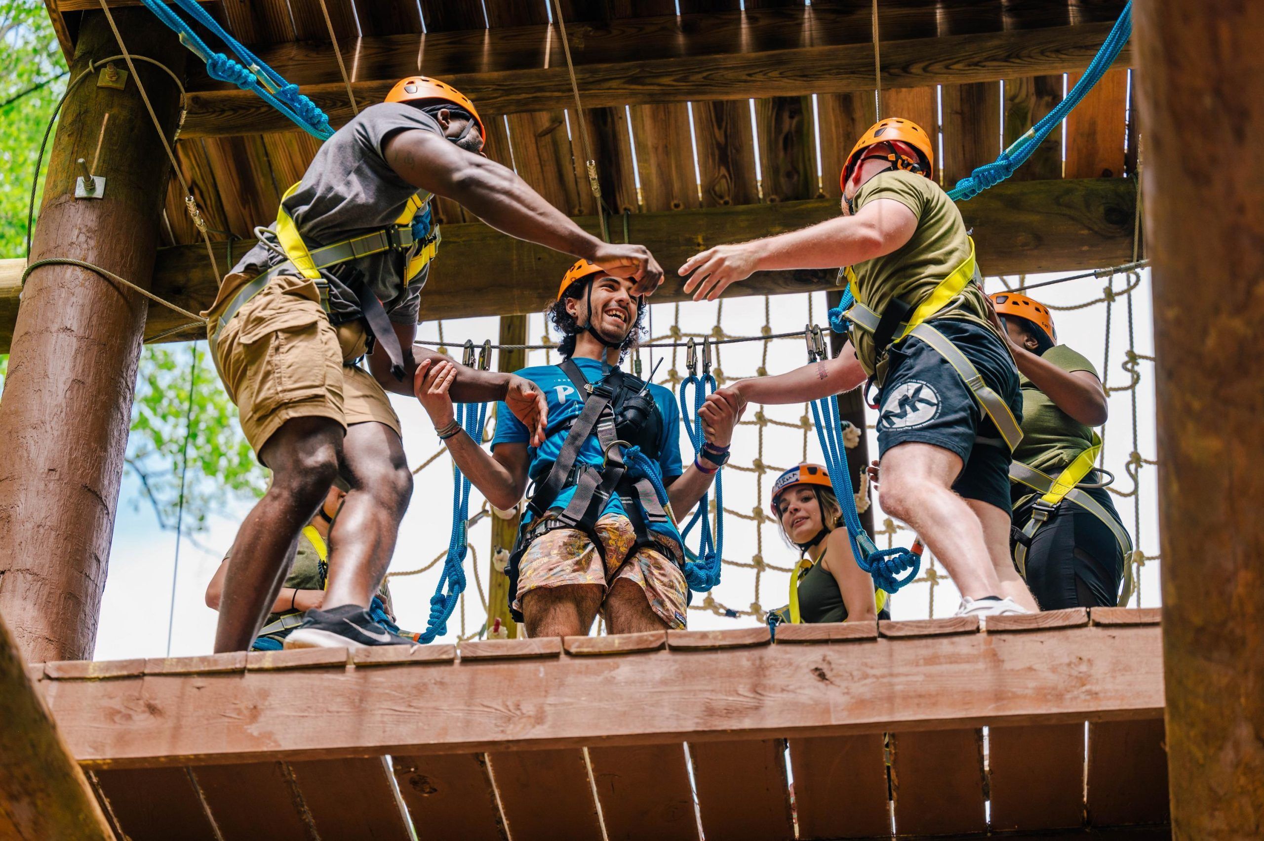 Several people wearing orange helmets and safety harnesses stand on a wooden platform during a ropes course activity. Blue climbing ropes are attached to the structure, and participants are assisting each other while preparing for the next challenge. The setting is outdoors with green foliage visible in the background.