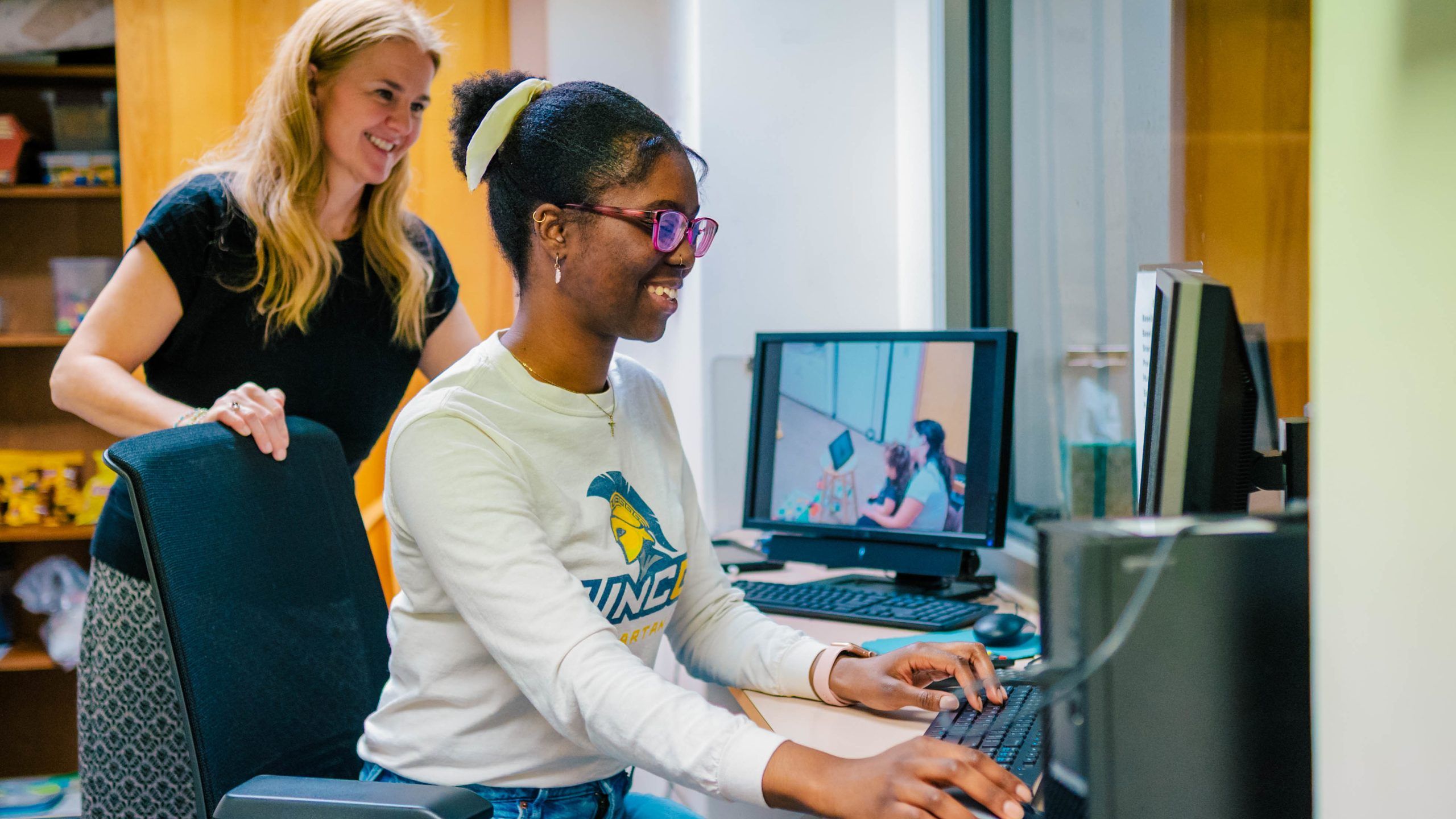 A person wearing a white long-sleeve shirt with a UNCG logo sits at a desk using a computer, typing on a keyboard. Another individual stands nearby, slightly behind the chair, observing. A monitor on the desk displays a video feed showing two people in conversation. The setting appears to be an office or lab space with shelves and equipment in the background.