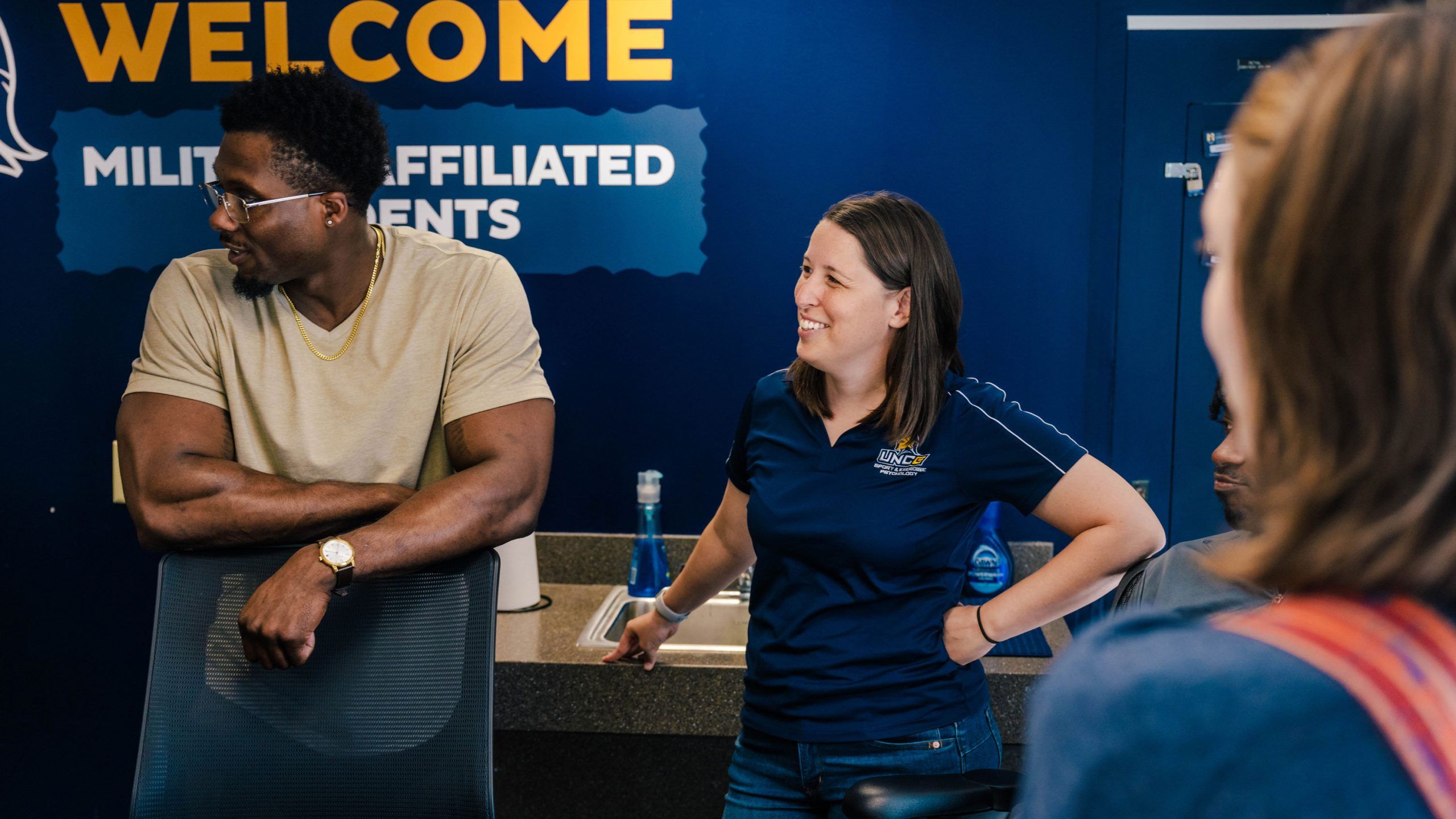 A group of people are gathered in a room with a dark blue wall featuring the words “WELCOME MILITARY AFFILIATED STUDENTS” in large yellow and white letters. One person is leaning on a chair wearing a light-colored T-shirt and a watch, while another stands nearby in a navy polo shirt with a university logo. A countertop with a water bottle and other small items is visible behind them, and part of another person is seen in the foreground wearing a striped shoulder strap. The setting appears to be an informal meeting or discussion area.
