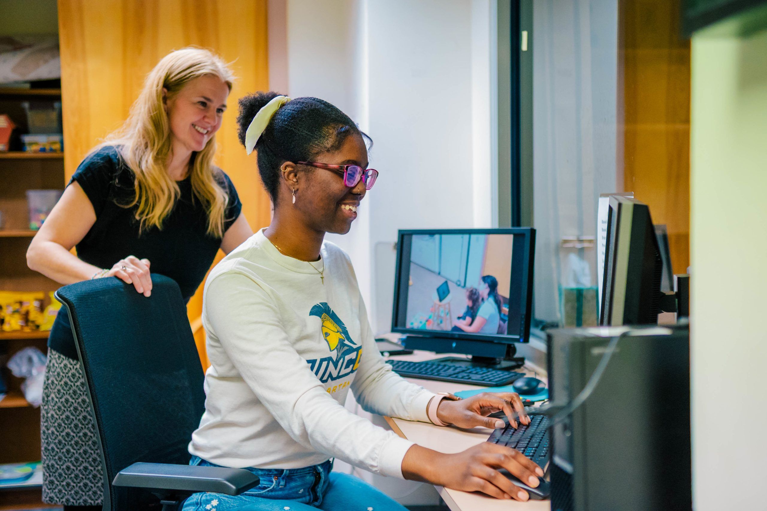 Undergrad Jahleen Gourdine and UNCG researcher Dr. Lenka Shriver in an office setting working at a computer; Jahleen is seated typing on a keyboard while Dr. Shriver stands nearby, with a monitor displaying a video feed of the same workspace.
