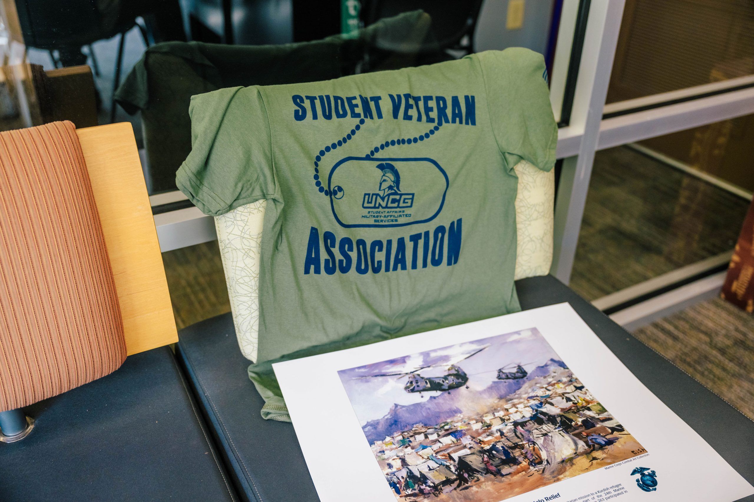 A green T-shirt with “Student Veteran Association” and the UNC Greensboro logo printed in blue is draped over a chair. In front of the chair is a colorful poster featuring military helicopters and soldiers in a combat scene. The setting appears to be indoors, near a glass wall and office furniture.
