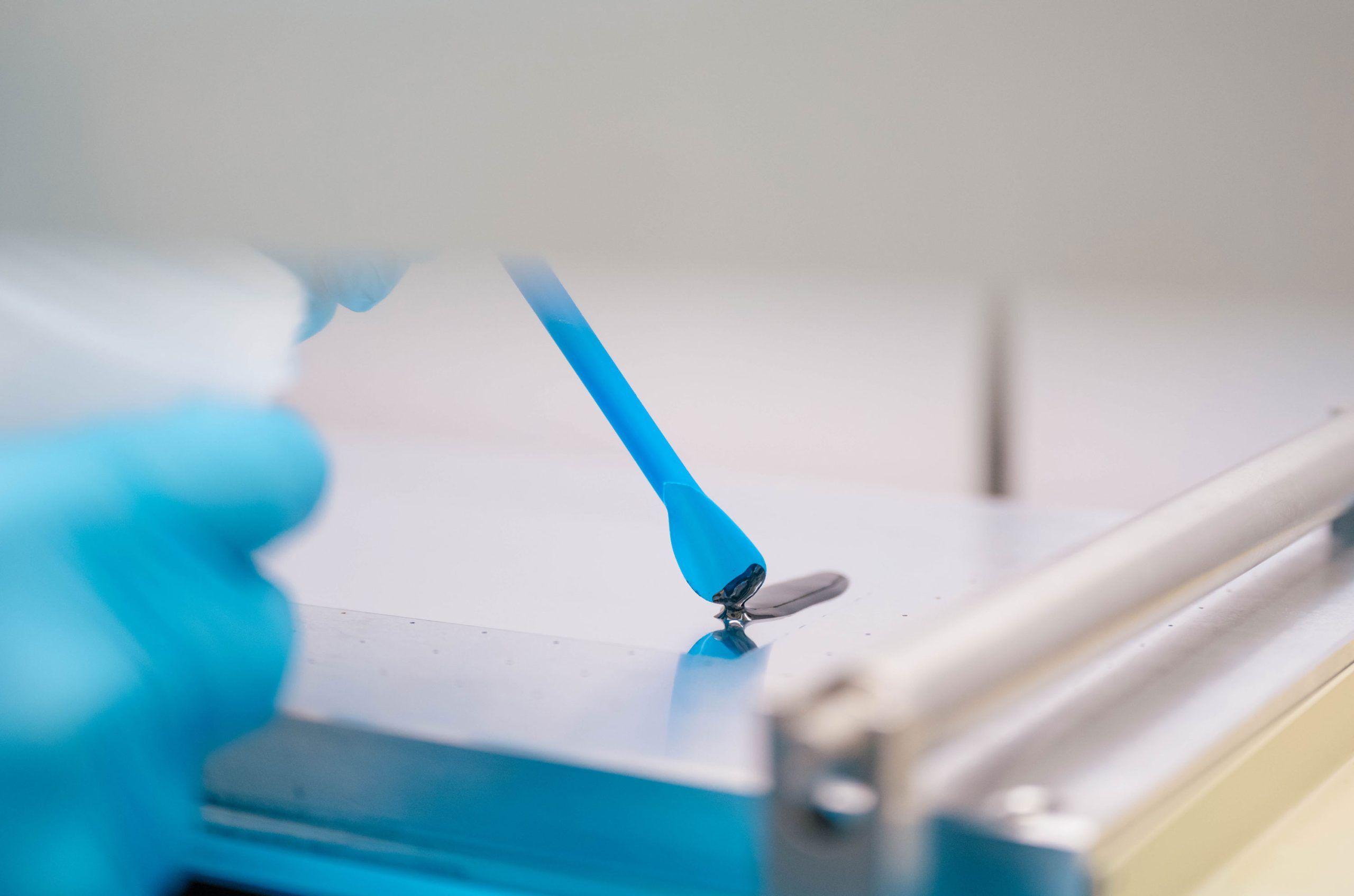 A close-up of a gloved hand using a blue spatula to spread or collect a small amount of dark material on a flat metallic surface. The setup appears to be part of a laboratory process, with clean, controlled conditions and precise handling of the substance.