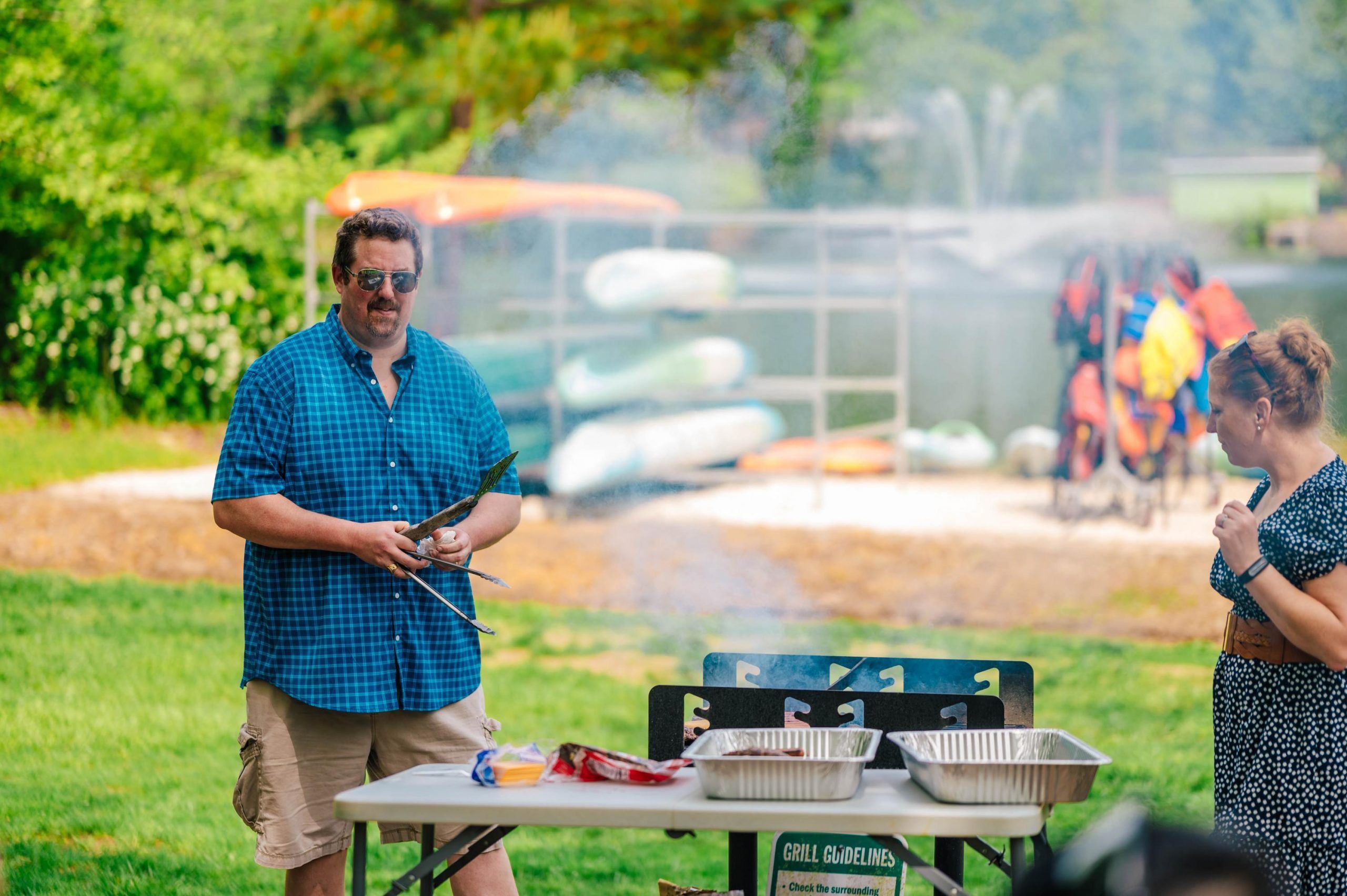 Two people stand outdoors near a grill with aluminum trays and cooking utensils on a table. One person is holding skewers, and the other is standing nearby. In the background, there are stacked kayaks, life jackets, and a lake with a fountain, suggesting a recreational area or picnic setting.