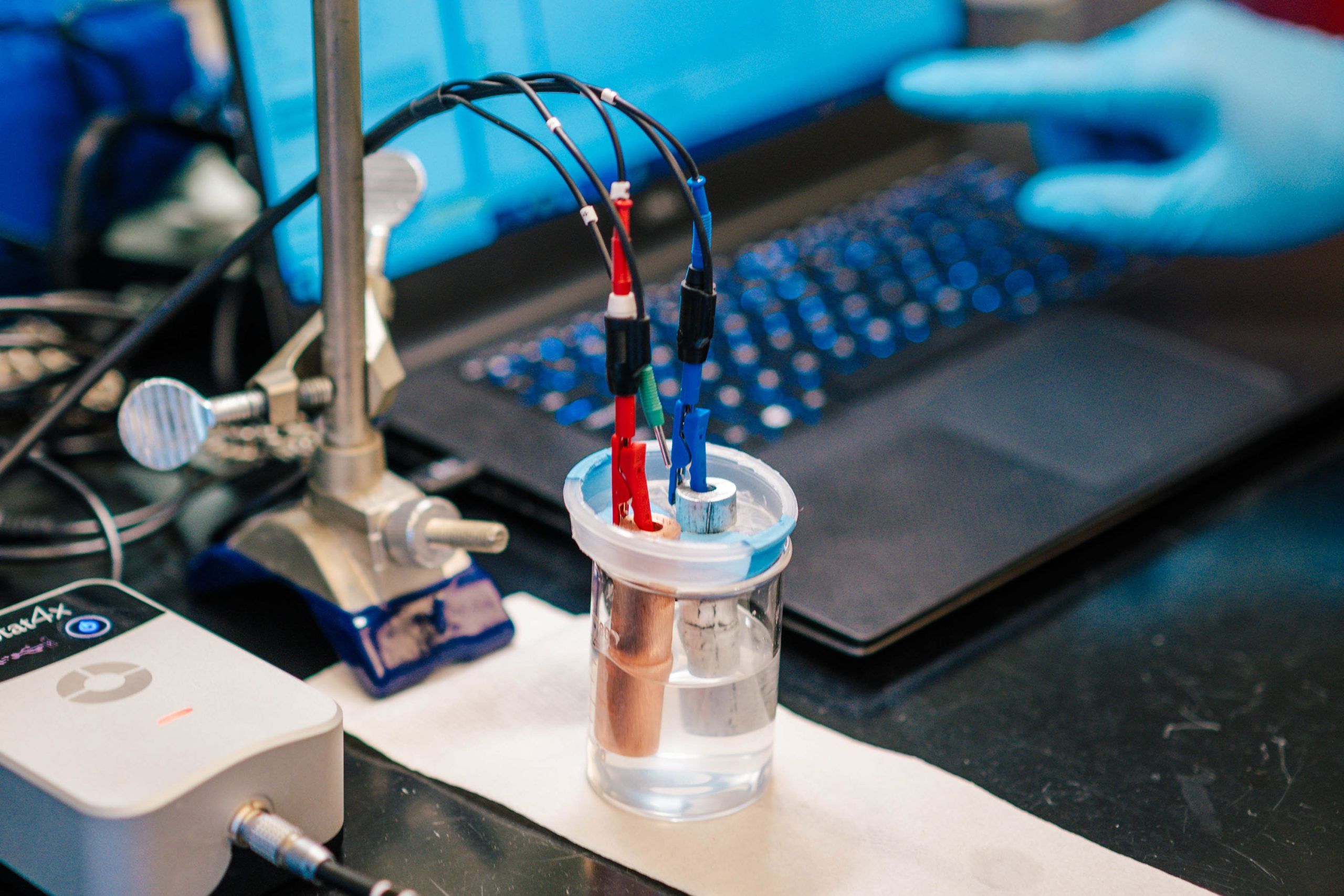 A laboratory setup showing an electrochemical experiment. A small clear container filled with liquid holds two metal electrodes connected to red, blue, and black wires. The wires are attached to clips and lead to a nearby device and a laptop in the background. A gloved hand is pointing toward the laptop screen, suggesting data monitoring or adjustment during the experiment.