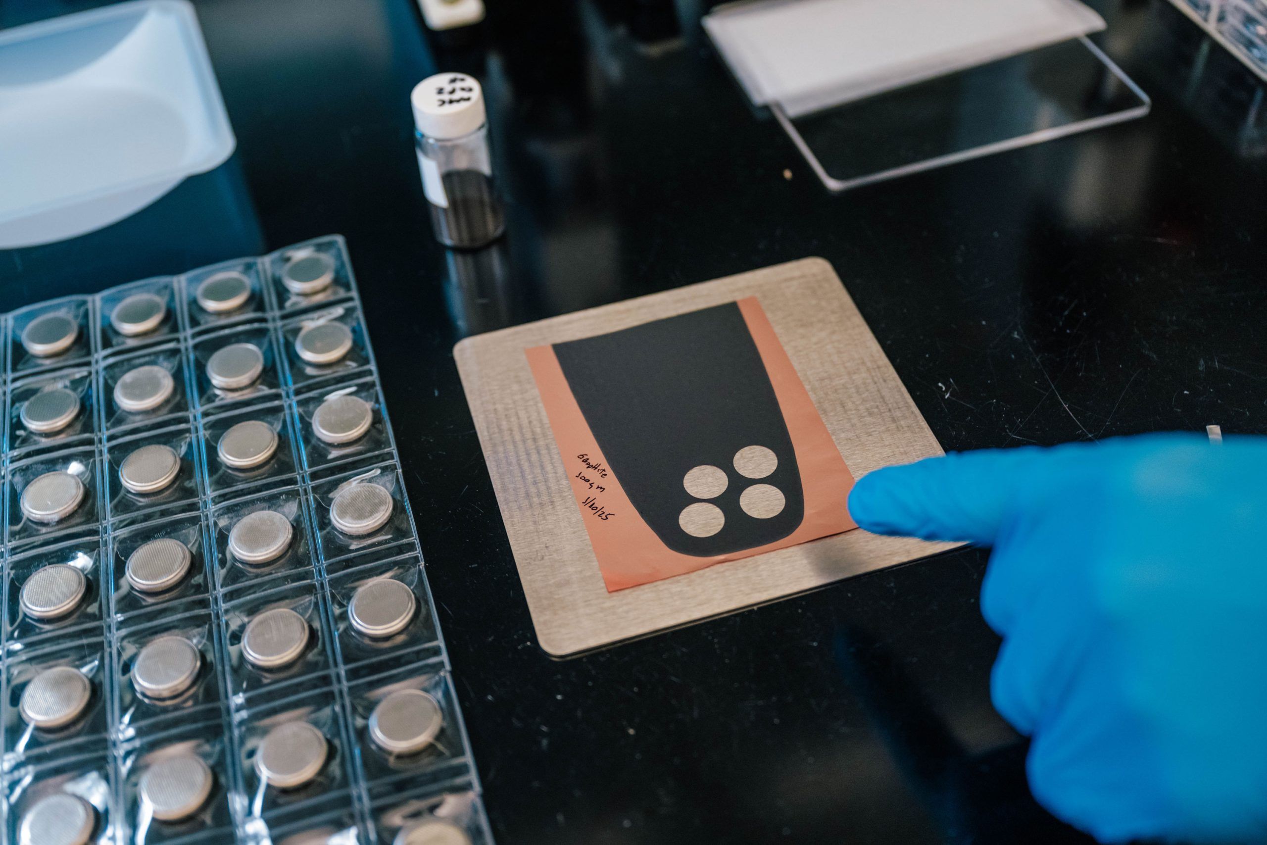 A gloved hand points to a square copper sheet with a black coating and three circular silver-colored samples placed on it. The sheet is labeled with handwritten text. Nearby, there is a tray holding multiple coin cell batteries, a small vial, and other lab materials on a black work surface, indicating a battery or materials science experiment.