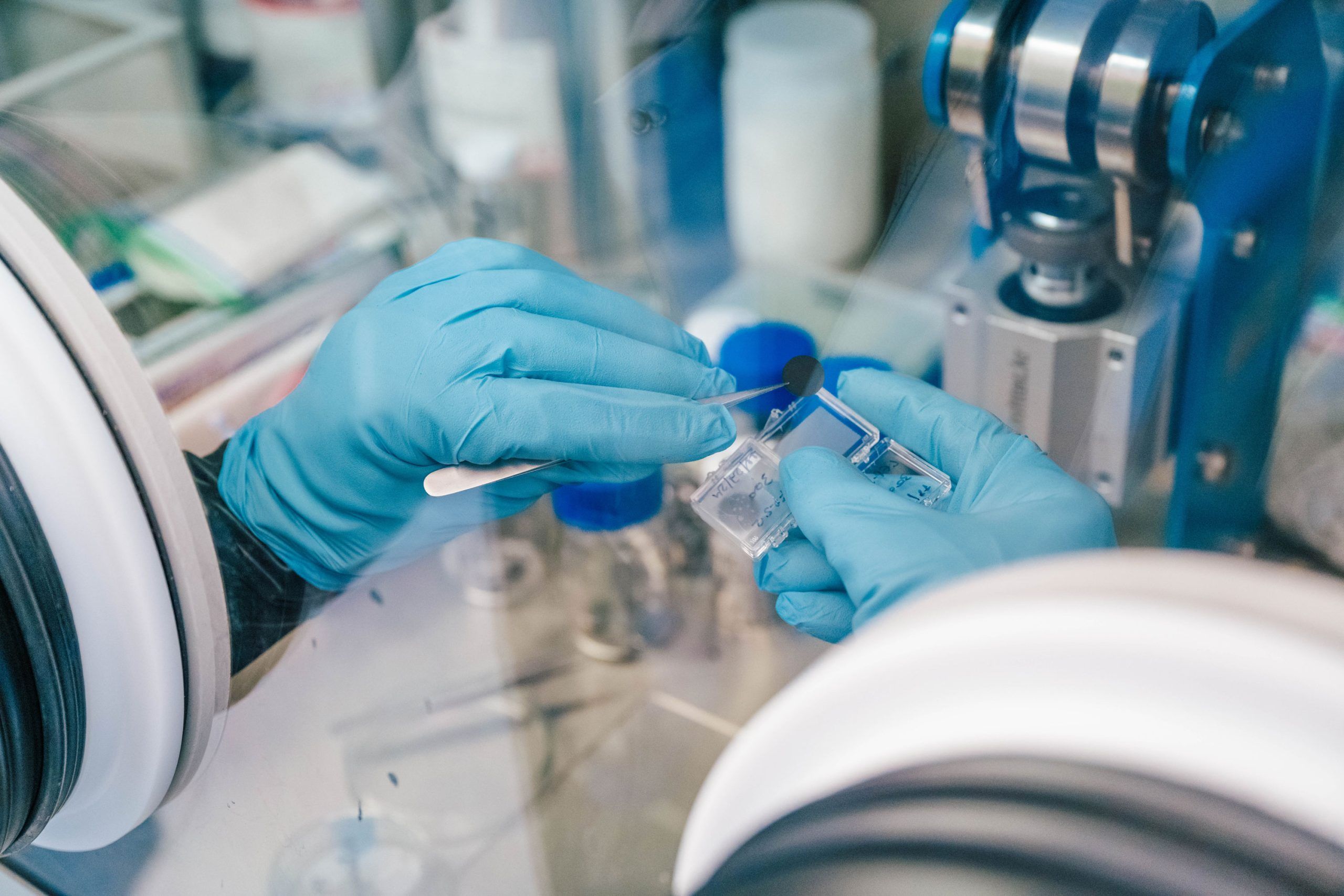 A close-up of two gloved hands working inside a laboratory glove box. One hand holds a small metal spatula, transferring a dark powder into a clear plastic container held by the other hand. The background shows lab equipment, including blue caps, a cylindrical component, and a mechanical arm, indicating a controlled environment for handling sensitive materials.