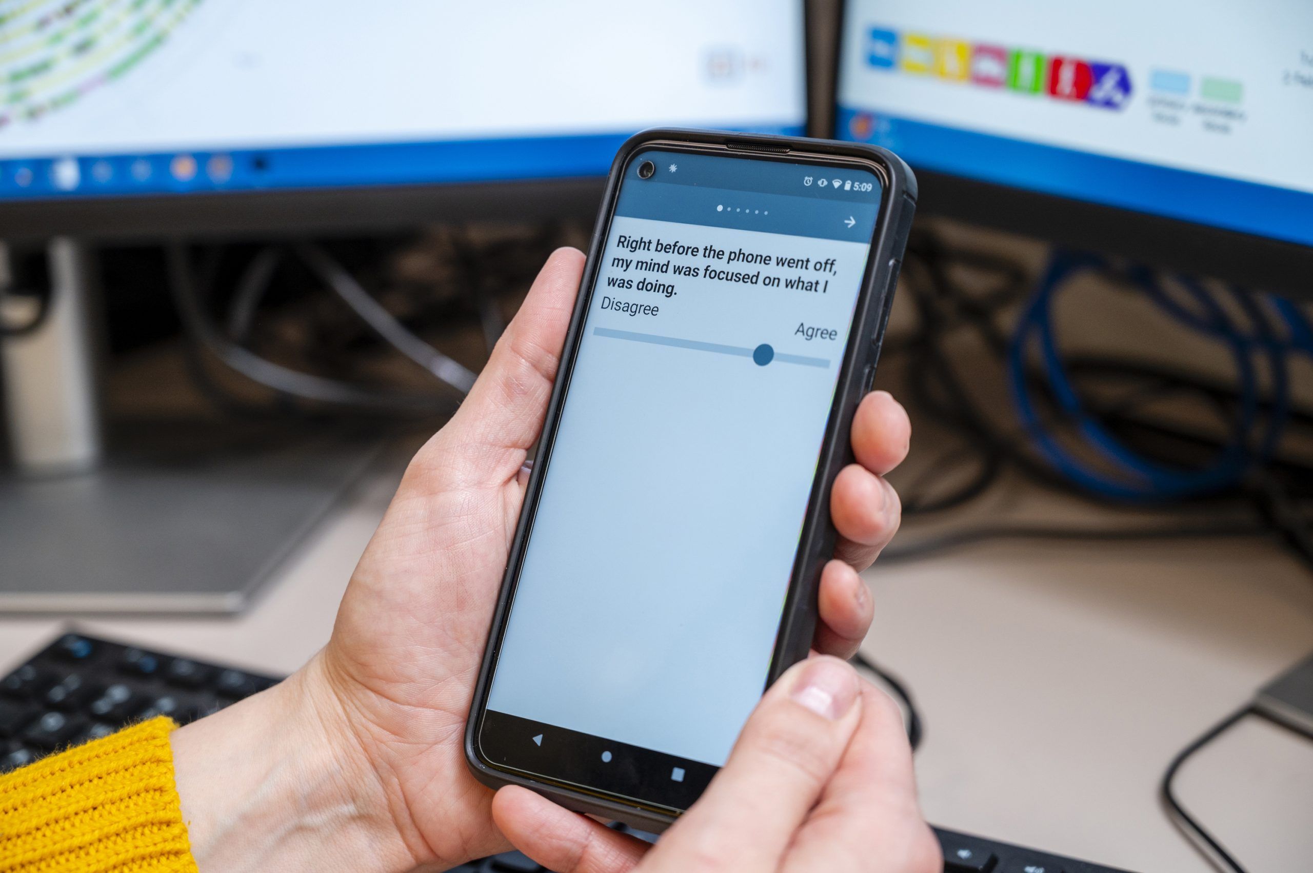 Dr. Maher holds a smartphone displaying a survey question that reads, “Right before the phone went off, my mind was focused on what I was doing.” Below the text is a horizontal slider positioned near the “Agree” end. A computer keyboard and monitor are visible in the background on a desk.