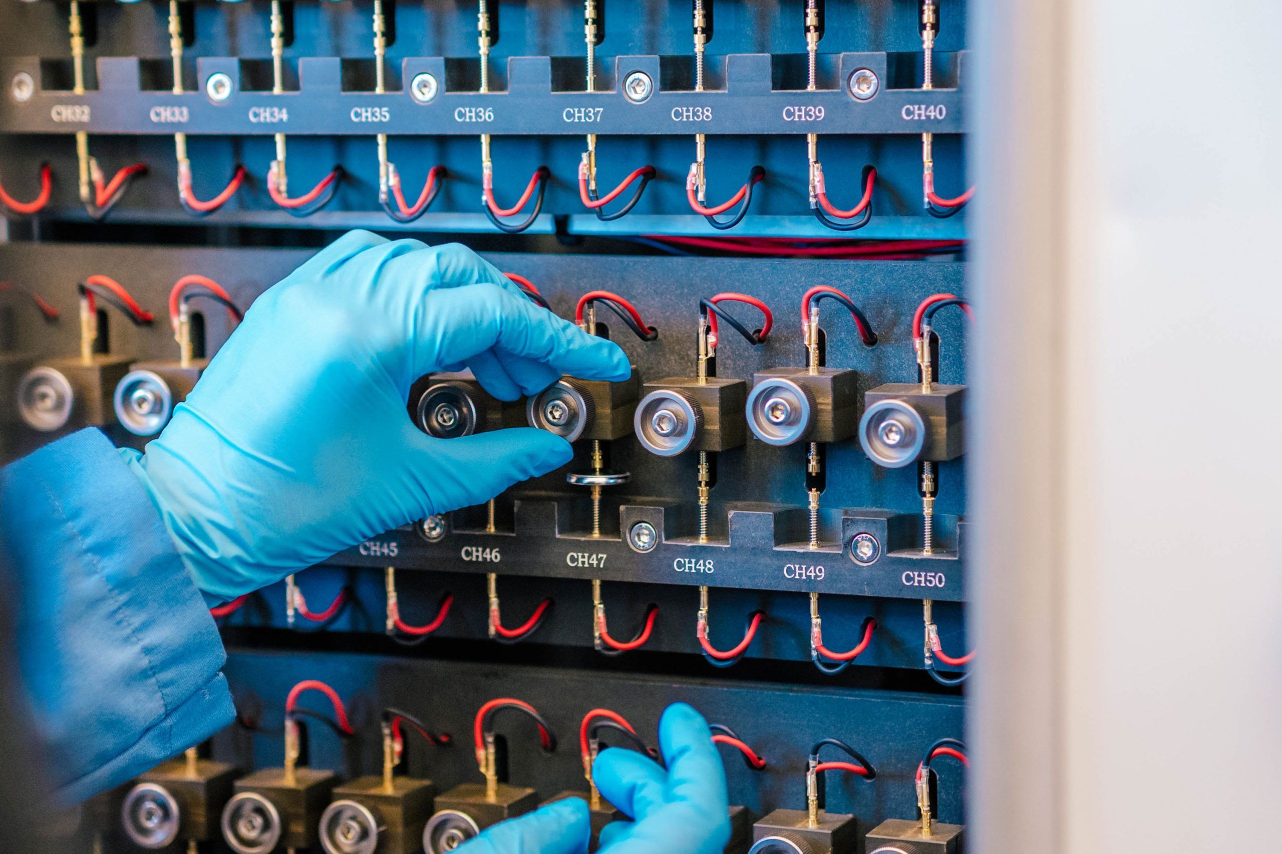 A close-up of gloved hands inserting or adjusting small cylindrical battery cells into a black testing rack with multiple labeled slots (CH33 to CH50). Each slot has red and black wires connected to the cells, indicating an electrical testing setup for batteries or energy storage components.