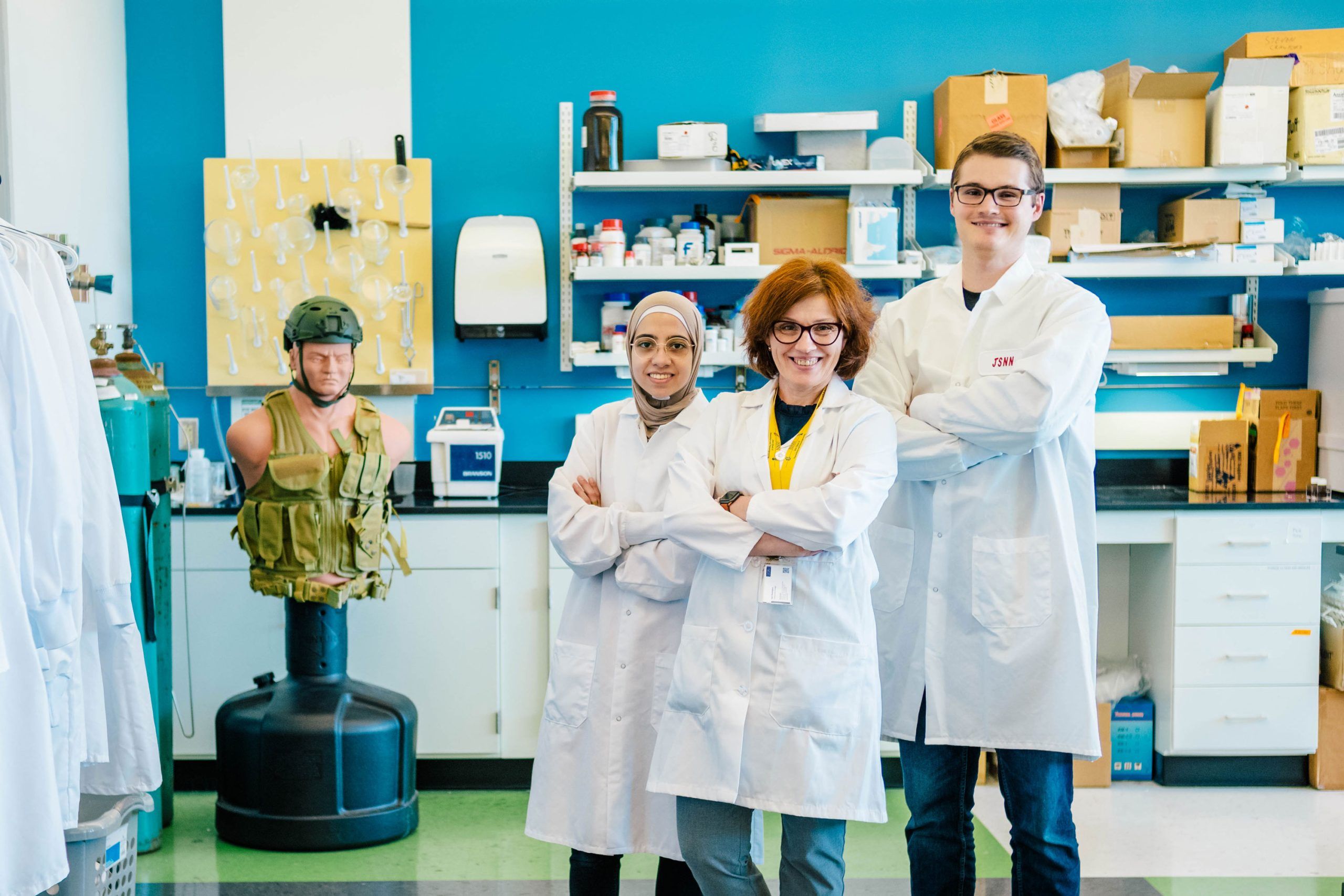 Three individuals wearing white lab coats stand confidently with arms crossed in a laboratory setting. Behind them are shelves filled with boxes, containers, and lab equipment. On the left side of the image, a mannequin torso is mounted on a stand and dressed in a green tactical vest, suggesting research related to protective gear. The background includes a bright blue wall and various scientific instruments on the counters.