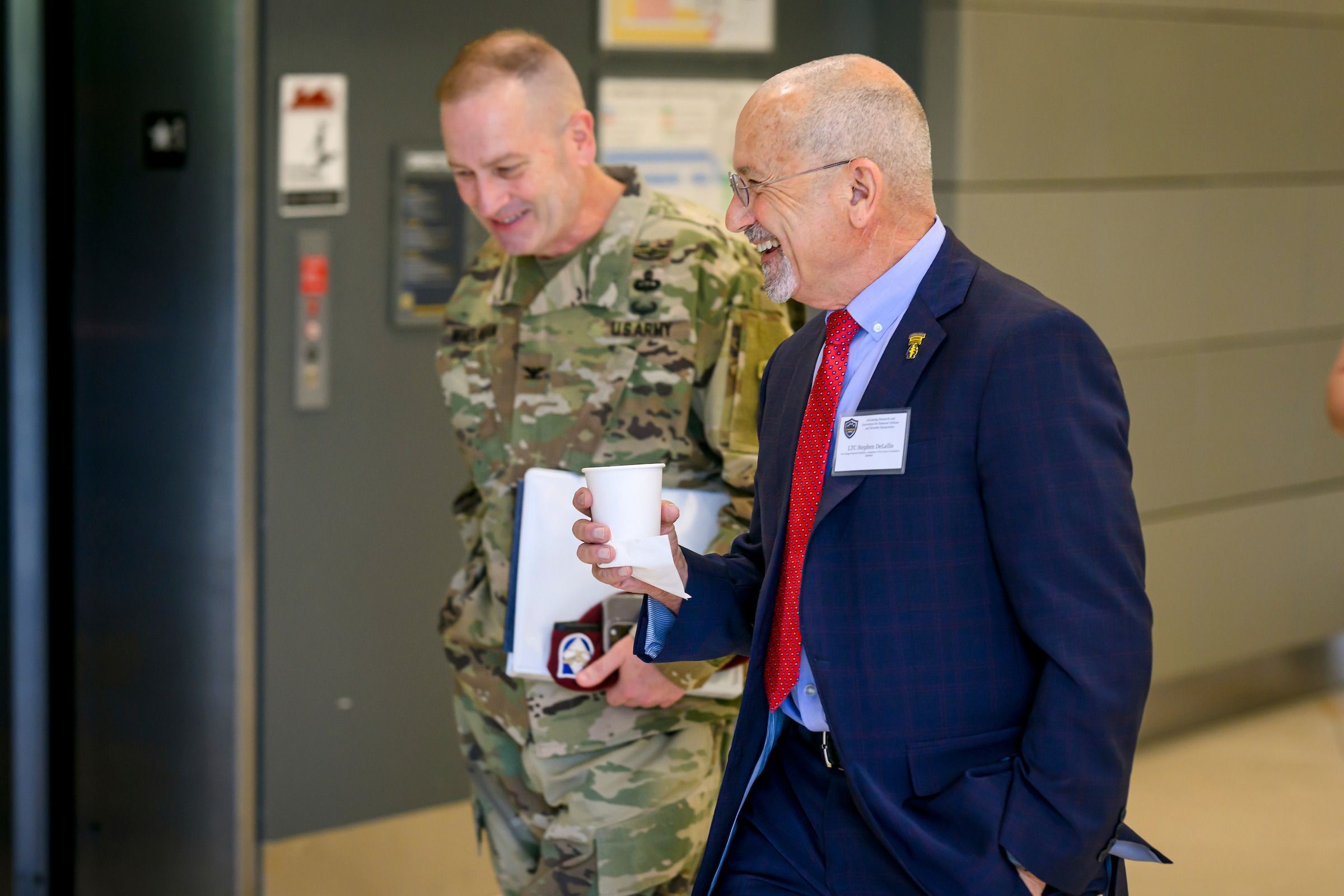 Two individuals walk through a hallway, one wearing a camouflage military uniform and holding a folder, the other dressed in a dark plaid suit with a red tie and carrying a paper cup. Behind them is an elevator door with signage and a gray wall, suggesting a professional or institutional setting.
