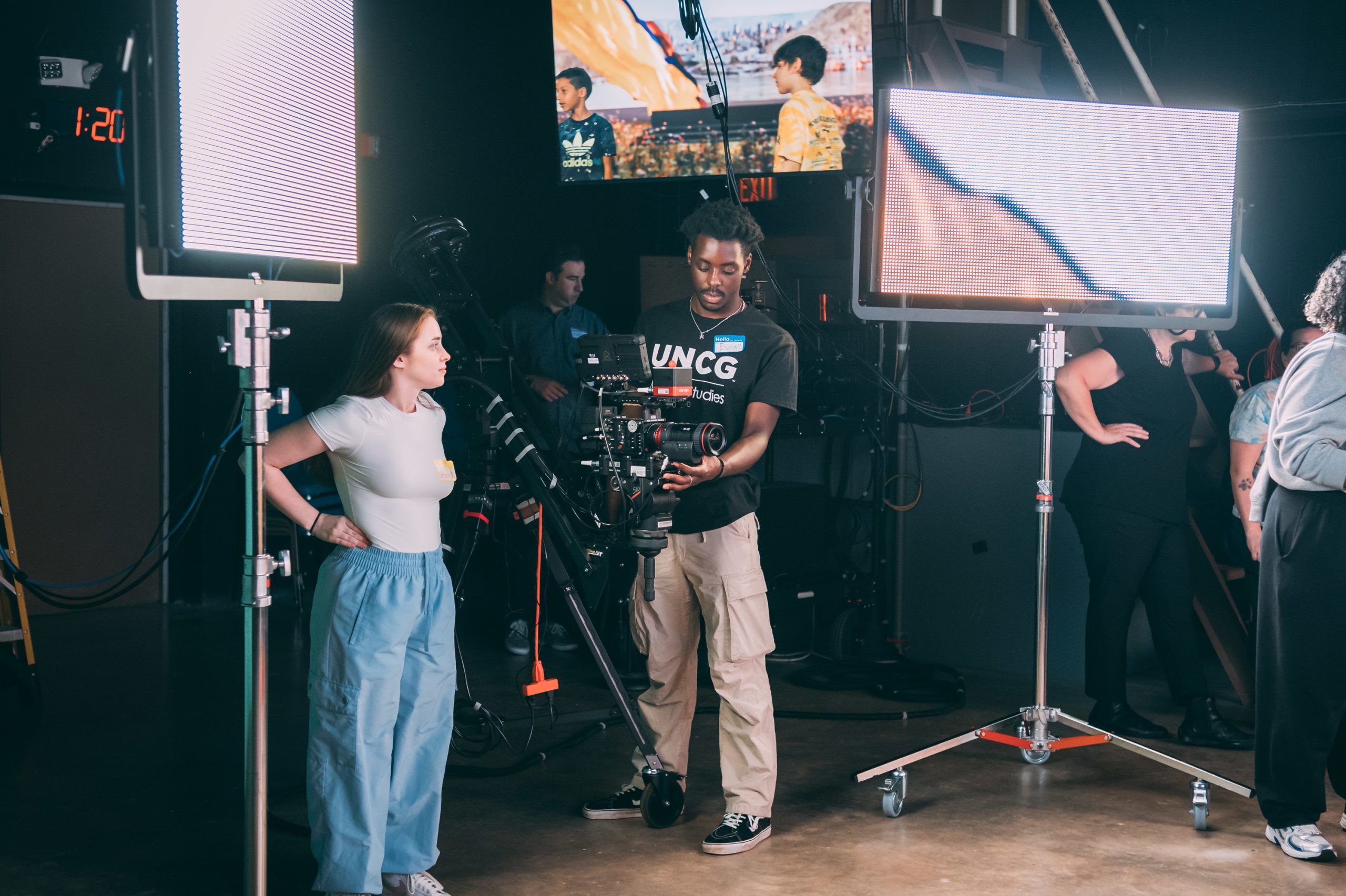 A film production setup with two large LED panel lights on stands and a person operating a professional camera rig mounted on a stabilizer. Another person stands nearby, appearing to discuss the shot. In the background, there are additional crew members, cables, and a monitor displaying a scene with two people outdoors. The environment is a dimly lit studio with equipment arranged for filming.