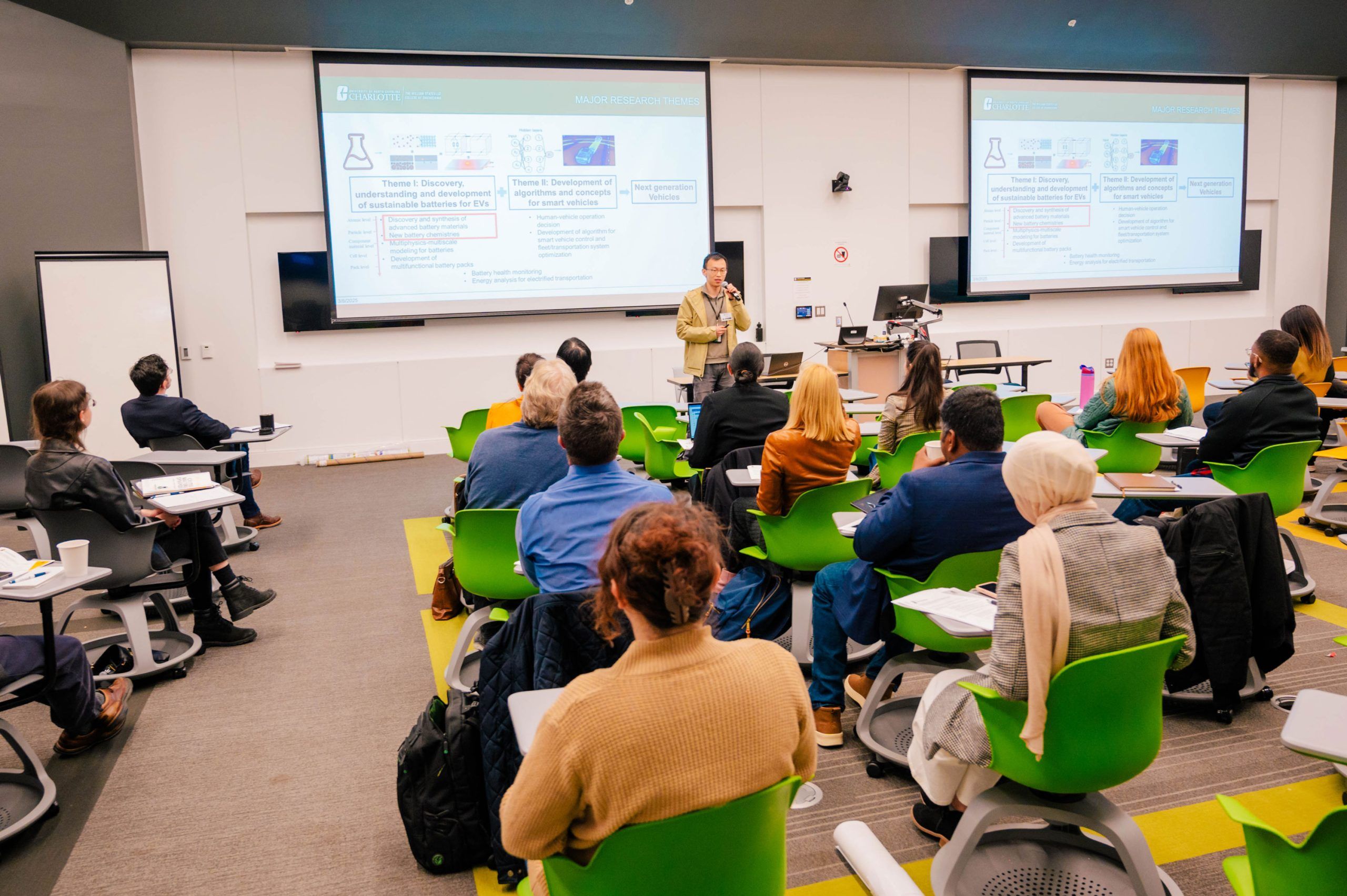 A classroom setting where a speaker stands at the front near a podium, presenting slides projected on two large screens. The slides contain diagrams, text, and headings related to scientific research topics. Attendees are seated in green chairs with attached desks, facing the presenter, and some have notebooks and papers. The room has bright lighting, white walls, and a modern design with multiple monitors and presentation equipment.