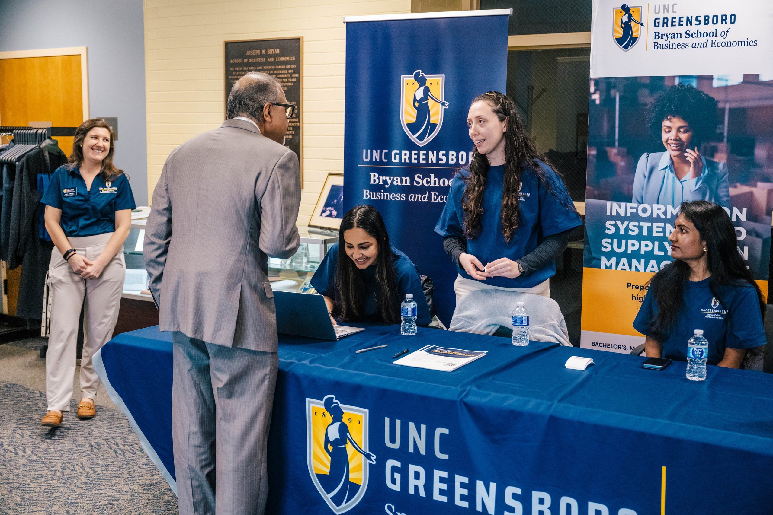 An information table at an event with a blue UNC Greensboro tablecloth and banners for the Bryan School of Business and Economics. Several people are seated or standing behind the table, which has water bottles, papers, and a laptop. A person in a suit is speaking to the representatives, and promotional signage about Information Systems and Supply Chain Management is visible in the background.